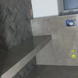 Szara łazienka z płytkami ułożonymi w jodełkę na ścianie, półka z płytek, otwory kanalizacyjne w ścianie, okno z szarą ramą.