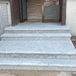 Granitowe schody wejściowe do budynku z metalową barierką, widok z dołu na drzwi wejściowe i otoczenie z zielenią.