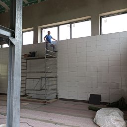 Artur Wolski usługi remontowo budowlane - Mężczyzna na rusztowaniu, w trakcie układania białych płytek na ścianie w pomieszczeniu z oknami, widoczne narzędzia i materiały budowlane, surowy stan wykończenia sufitu i podłogi.