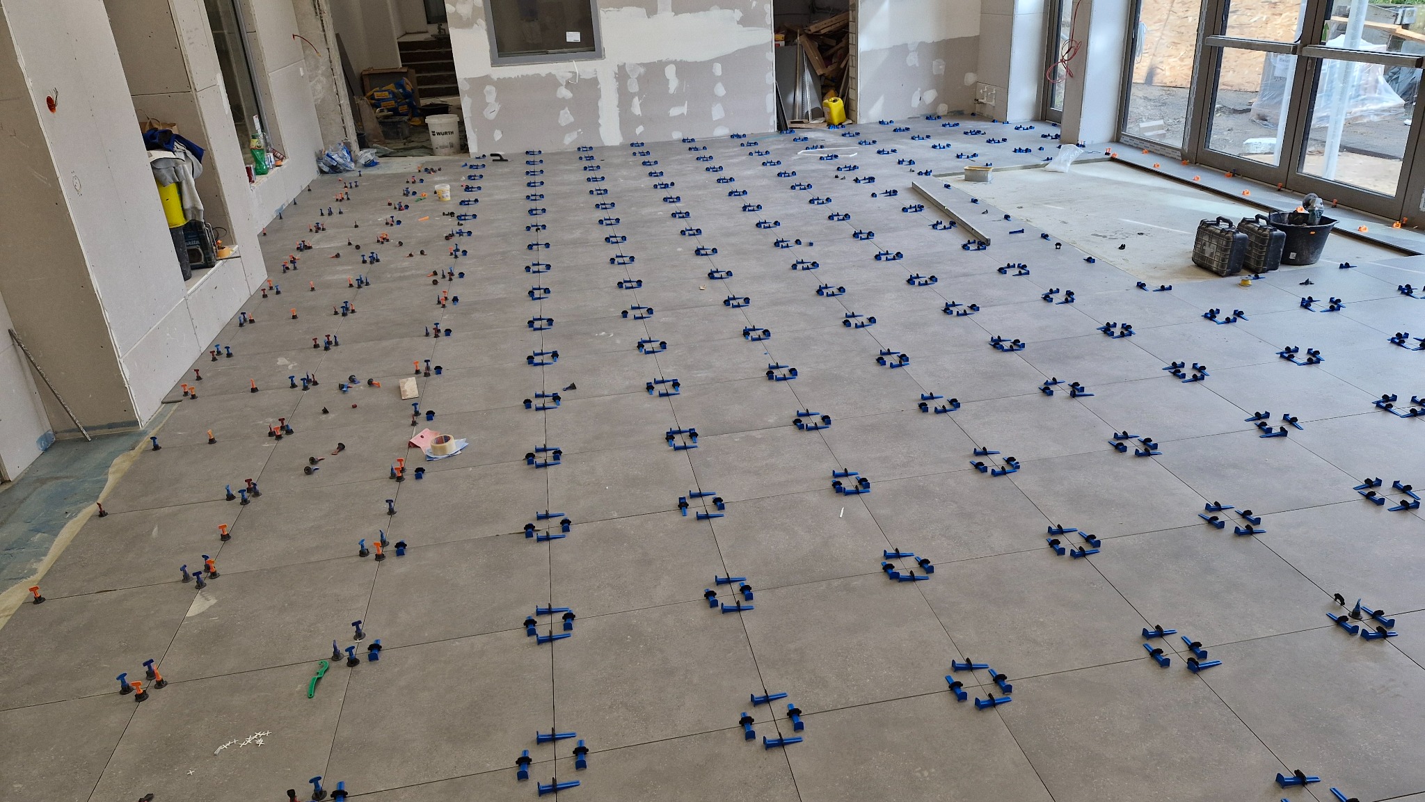 Szare płytki podłogowe w trakcie układania, z użyciem niebieskich i pomarańczowych klipsów poziomujących, widoczne ślady po zaprawie, w tle surowe ściany w trakcie wykończenia.