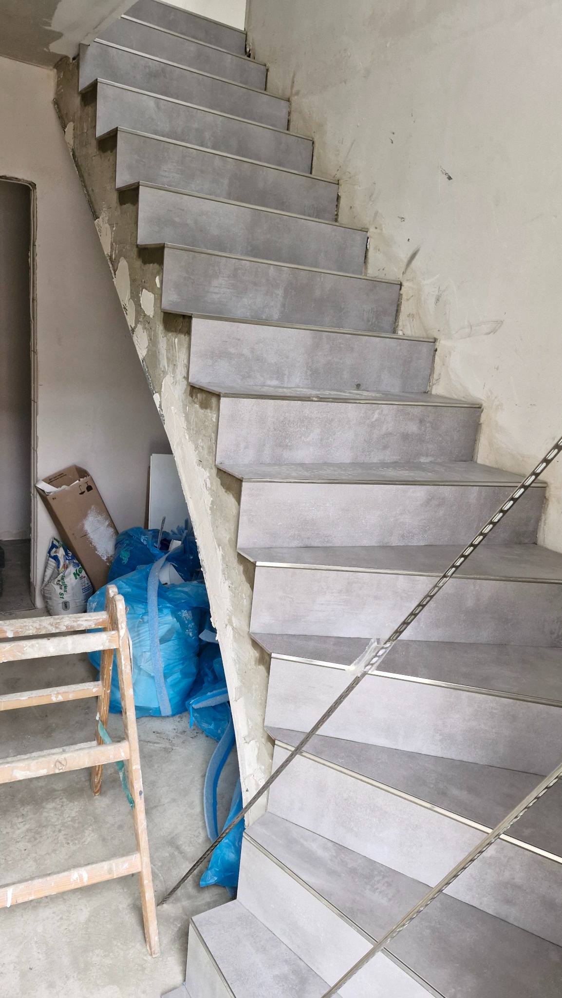 Schody w trakcie wykańczania, szare płytki z metalowymi listwami, widoczne worki z materiałami budowlanymi i drabina.