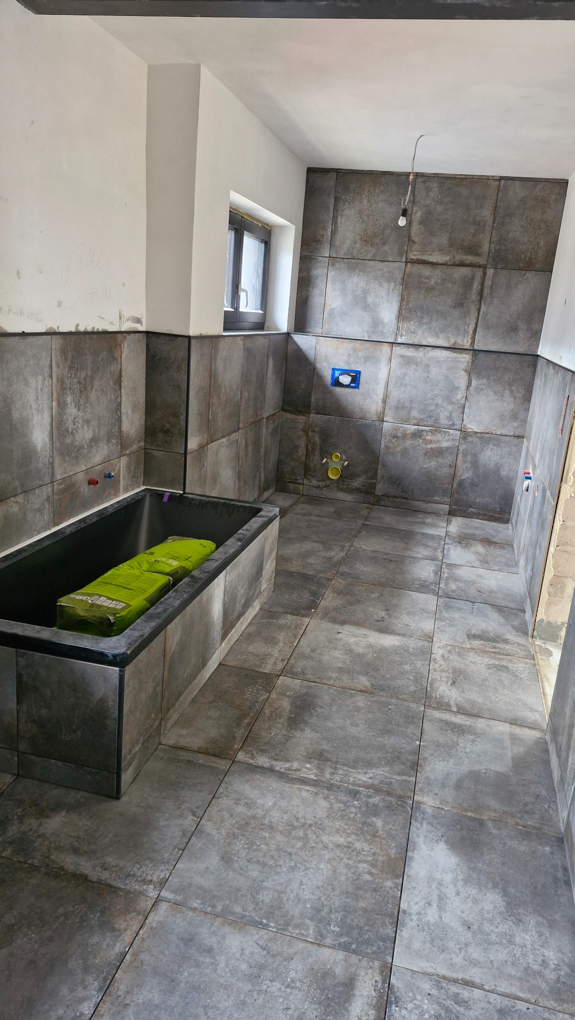 Łazienka w trakcie remontu z szarymi, kwadratowymi płytkami na ścianach i podłodze; czarna wanna wypełniona workami z zaprawą, okno w tle.