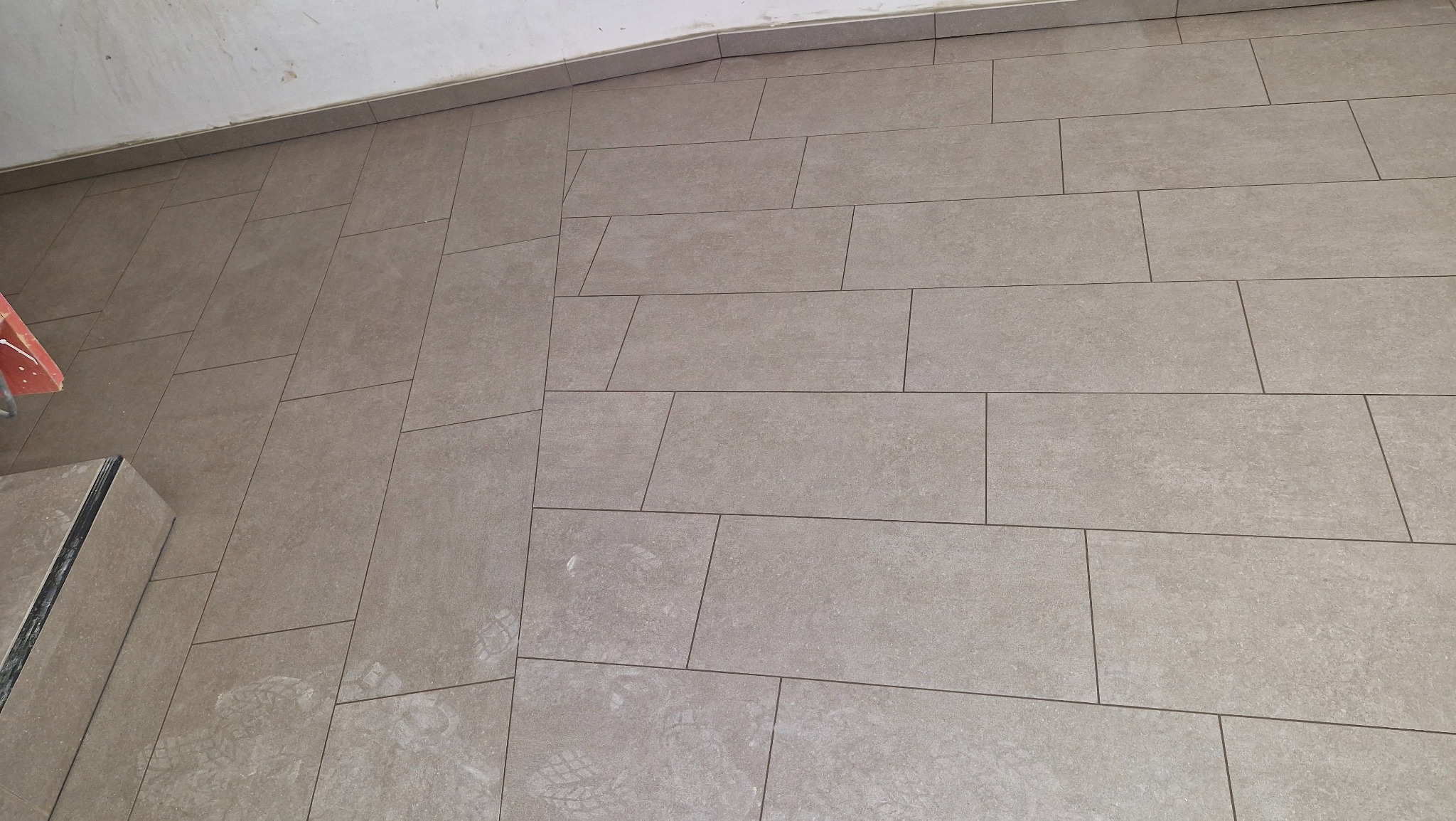 Ułożona podłoga z prostokątnych, beżowych płytek ceramicznych z ciemniejszą fugą, widoczne ślady stóp i fragment schodka z wykończeniem.