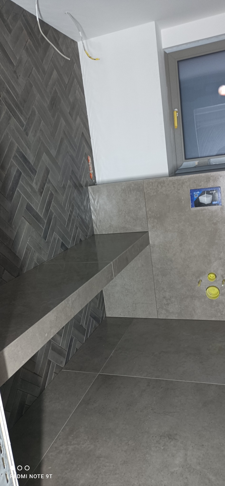 Szara łazienka z płytkami ułożonymi w jodełkę na ścianie, półka z płytek, otwory kanalizacyjne w ścianie, okno z szarą ramą.