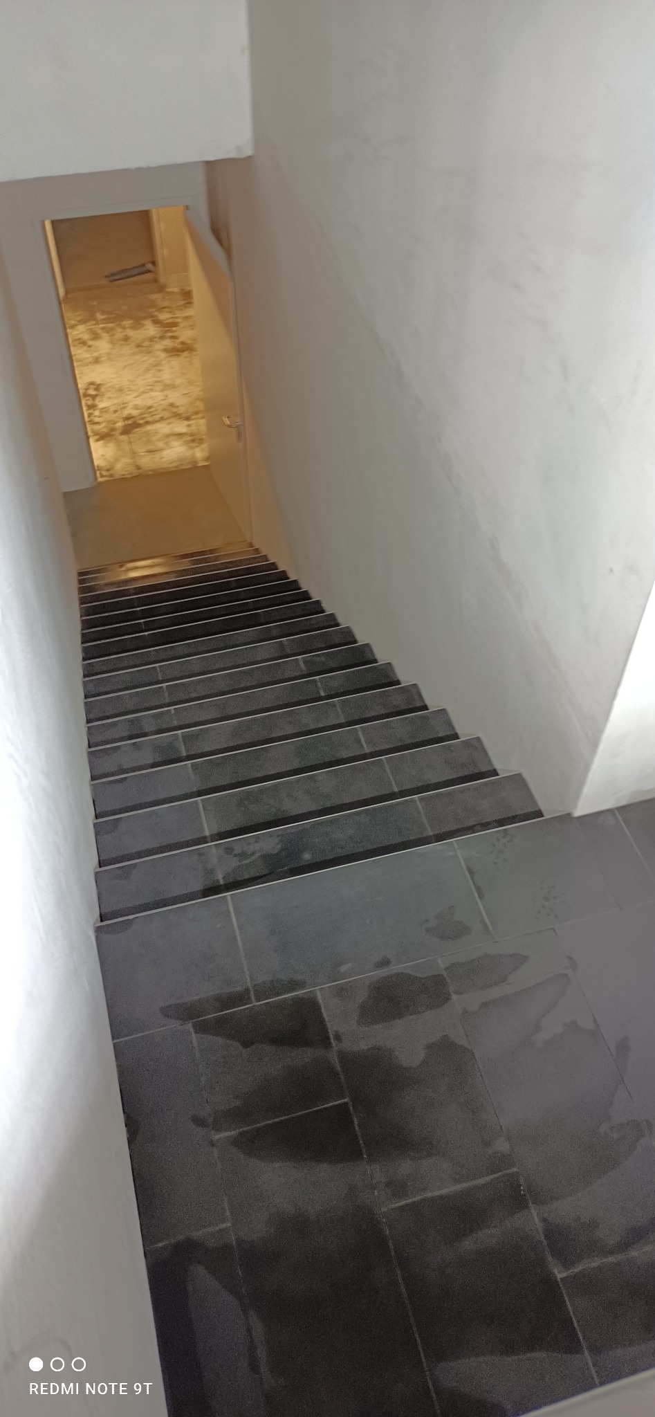 Schody wyłożone ciemnymi, błyszczącymi płytkami ceramicznymi, widoczne ślady po fugowaniu, ściany pomalowane na biało.