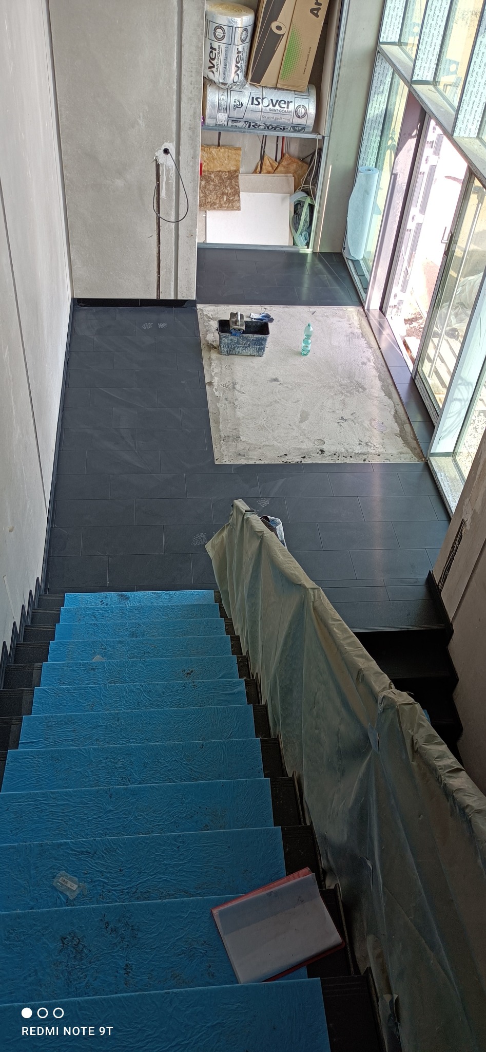 Montaż ciemnoszarych płytek na podłodze w korytarzu, widok z góry na zabezpieczone folią schody, pojemnik z narzędziami i butelki z wodą na niedokończonej powierzchni.