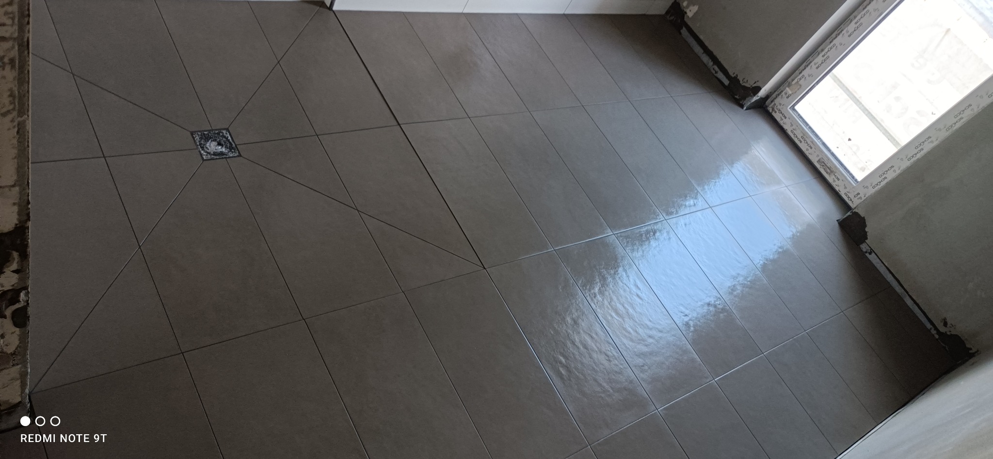 Ułożone, błyszczące, szare płytki podłogowe w łazience, z odpływem liniowym w kształcie kwadratu w rogu i oknem w tle, widoczne ślady fugowania przy ścianach.