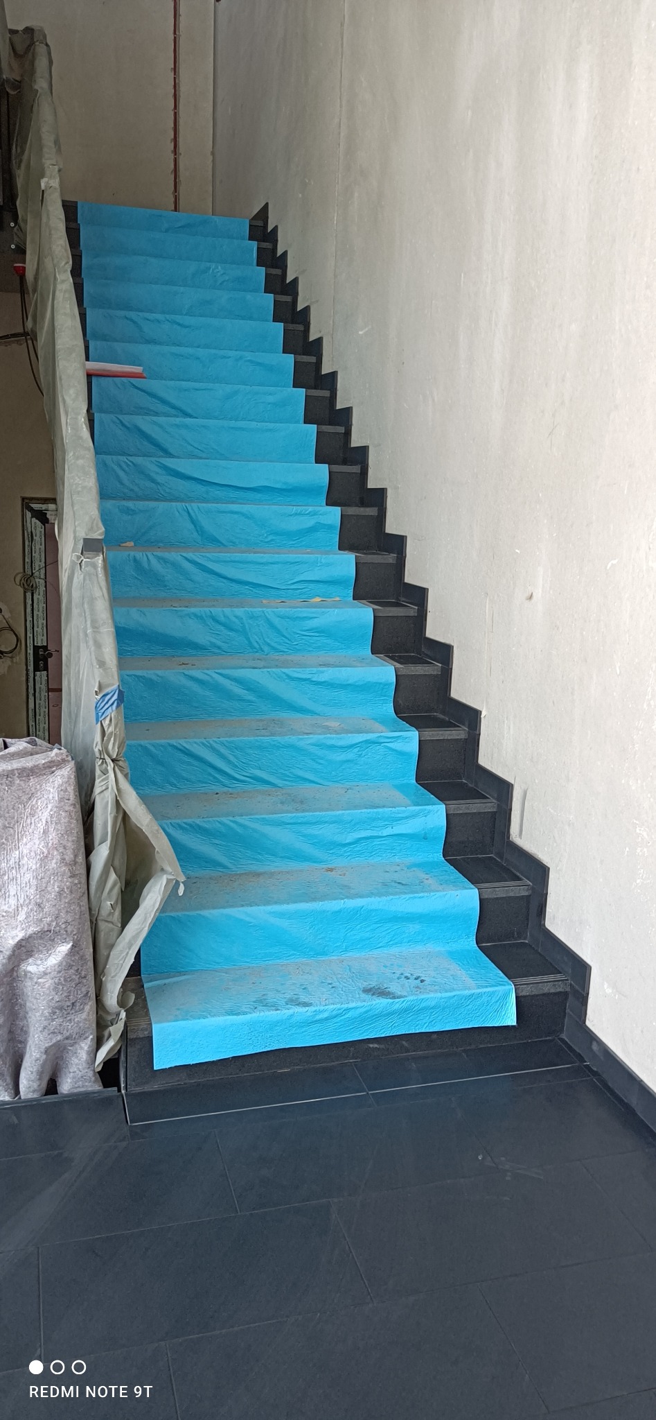 Schody z ciemnego kamienia zabezpieczone niebieską folią ochronną podczas remontu, widoczne zakryte folią poręcze i fragment ściany w surowym stanie.