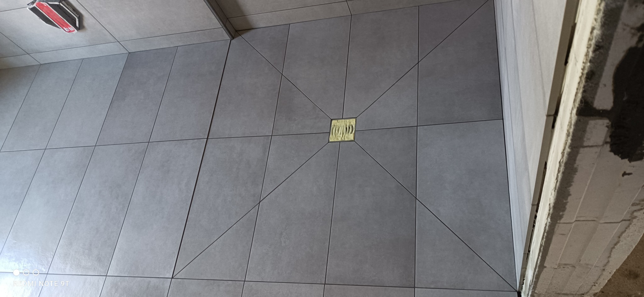 Szare, kwadratowe płytki podłogowe ułożone ze złotym odpływem liniowym pośrodku, widoczne elementy surowego wykończenia ściany i narzędzie glazurnicze w rogu.