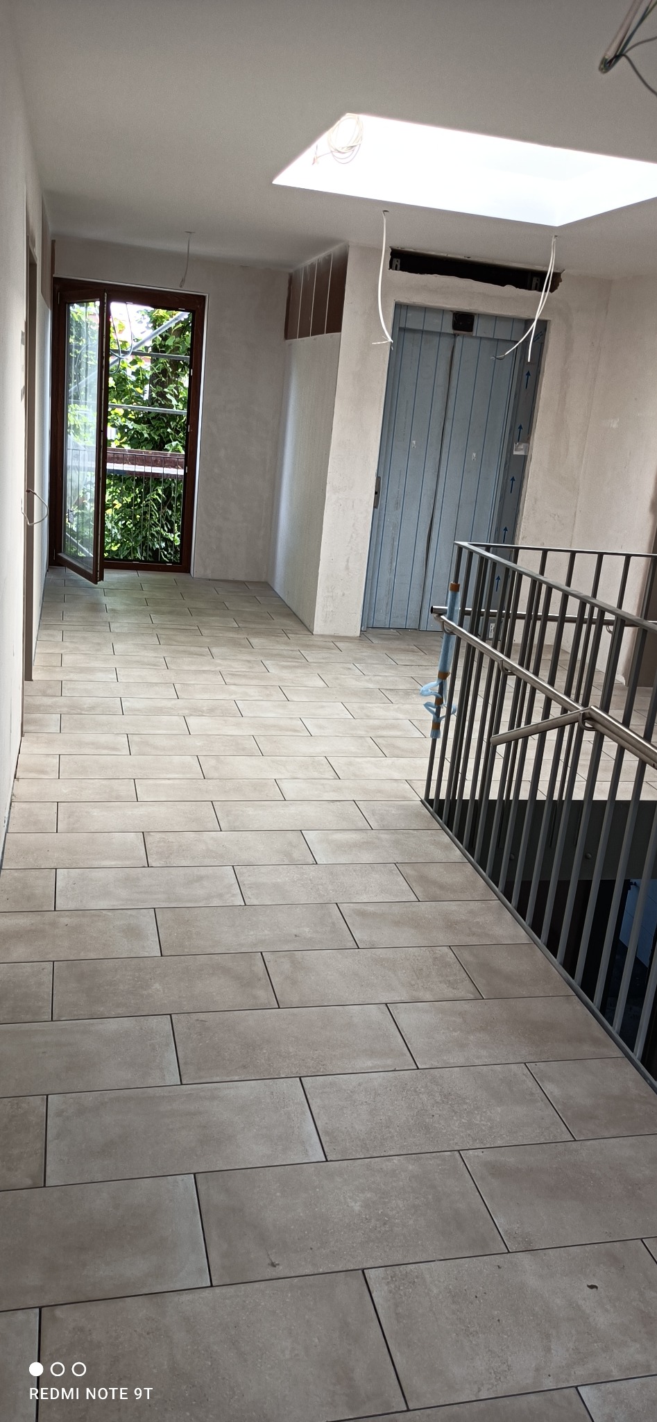 Korytarz w trakcie remontu z położoną beżową podłogą z płytek, otwartymi drzwiami balkonowymi i widocznym fragmentem schodów z metalową barierką. Na suficie widoczne okno dachowe i wiszące kable.