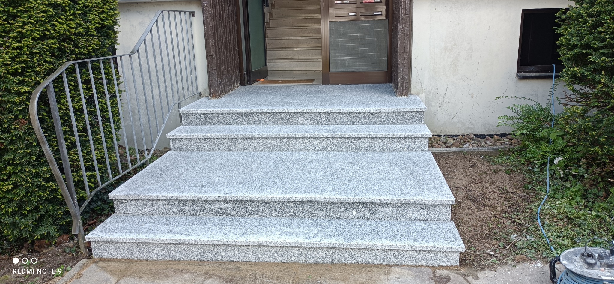 Granitowe schody wejściowe do budynku z metalową barierką, widok z dołu na drzwi wejściowe i otoczenie z zielenią.