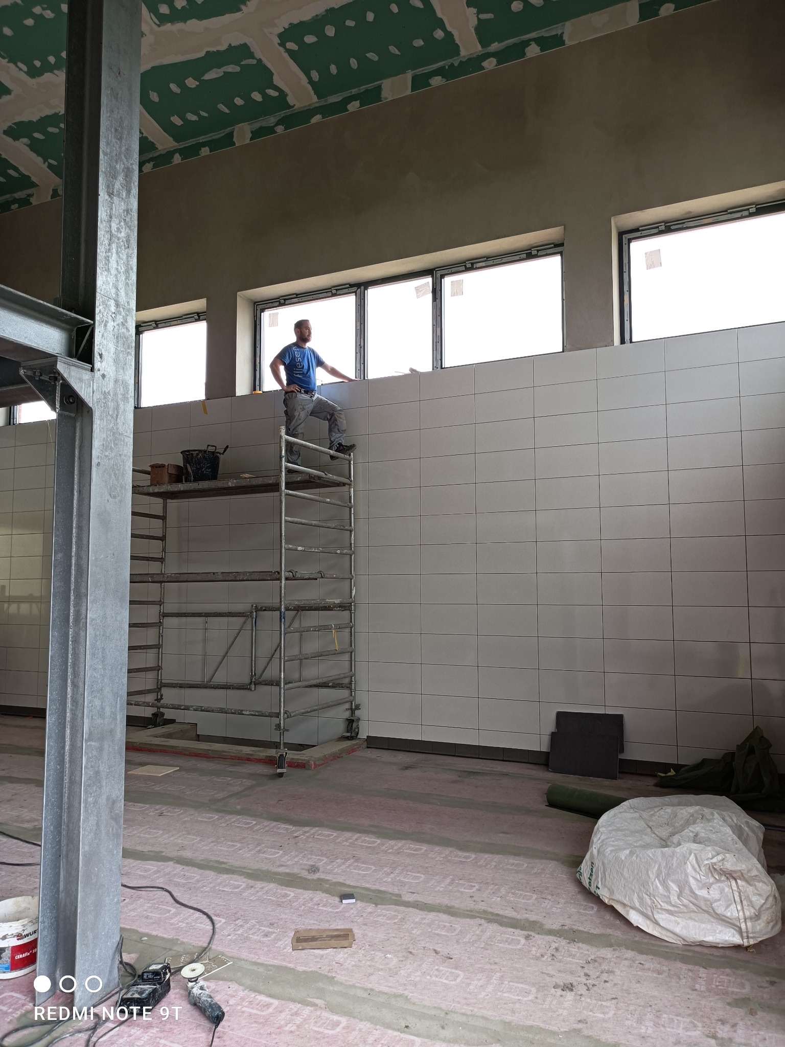 Mężczyzna na rusztowaniu, w trakcie układania białych płytek na ścianie w pomieszczeniu z oknami, widoczne narzędzia i materiały budowlane, surowy stan wykończenia sufitu i podłogi.