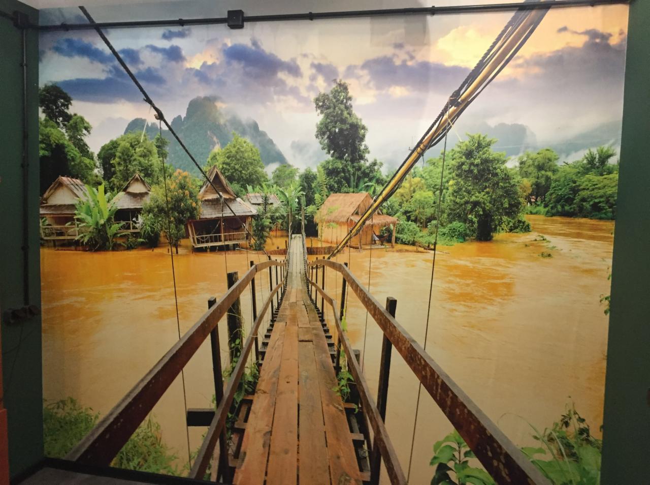 Fototapeta na ścianie przedstawiająca drewniany most linowy prowadzący do domków na palach w tropikalnym krajobrazie z rzeką o brązowej wodzie i górami w tle.