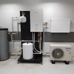 Zestaw instalacji grzewczej z pompą ciepła Panasonic Aquarea, zasobnikiem ciepłej wody i zbiornikiem buforowym, zamontowany na wózku transportowym w pomieszczeniu technicznym.