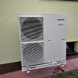 Zewnętrzna jednostka klimatyzacji Panasonic Inverter, zamontowana na białej, metalowej konstrukcji wsporczej przy ścianie budynku o jasnej elewacji, widoczne okno piwniczne i fragment okna...