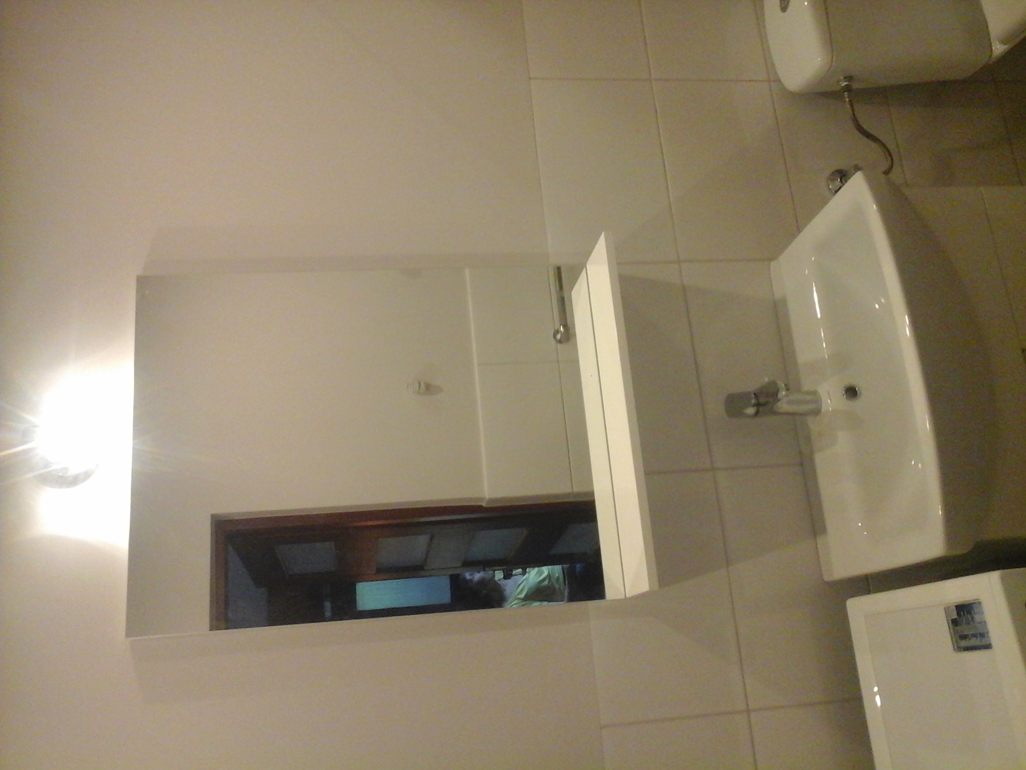 Umywalka wisząca na ścianie wyłożonej jasnymi, kwadratowymi płytkami, z widocznym lustrem szafki, w którym odbija się fragment innego pomieszczenia.