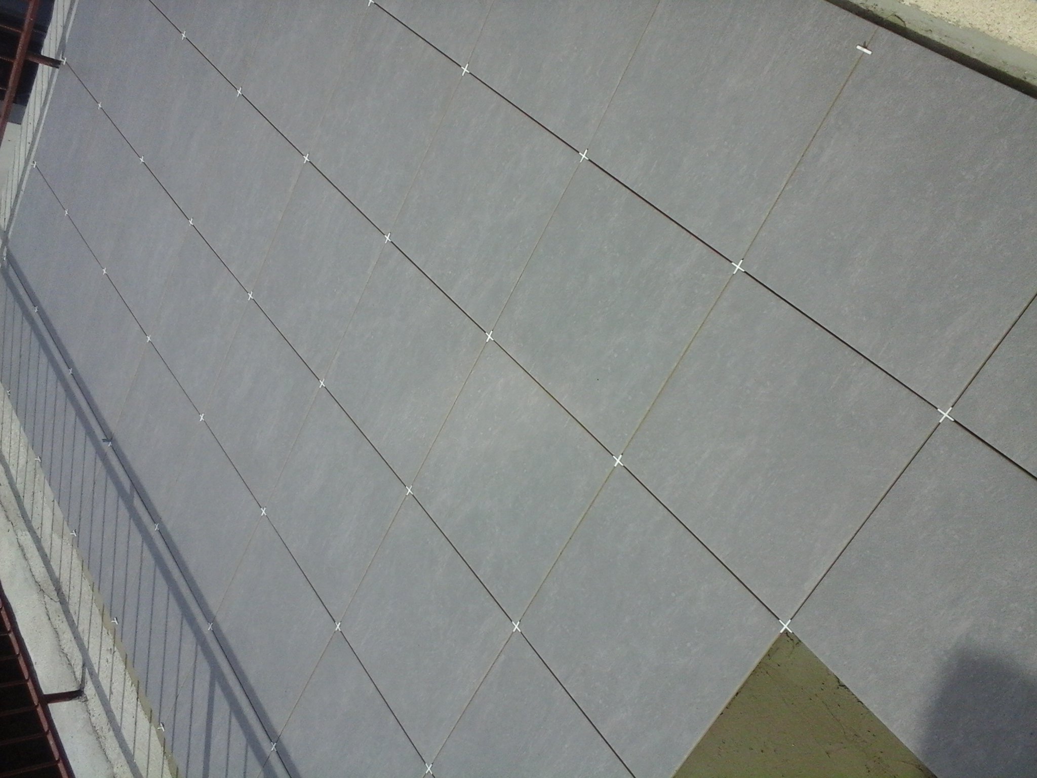 Szare płytki ceramiczne ułożone na ścianie zewnętrznej budynku, z widocznymi krzyżykami dystansowymi w spoinach, fragment balkonu po lewej stronie.