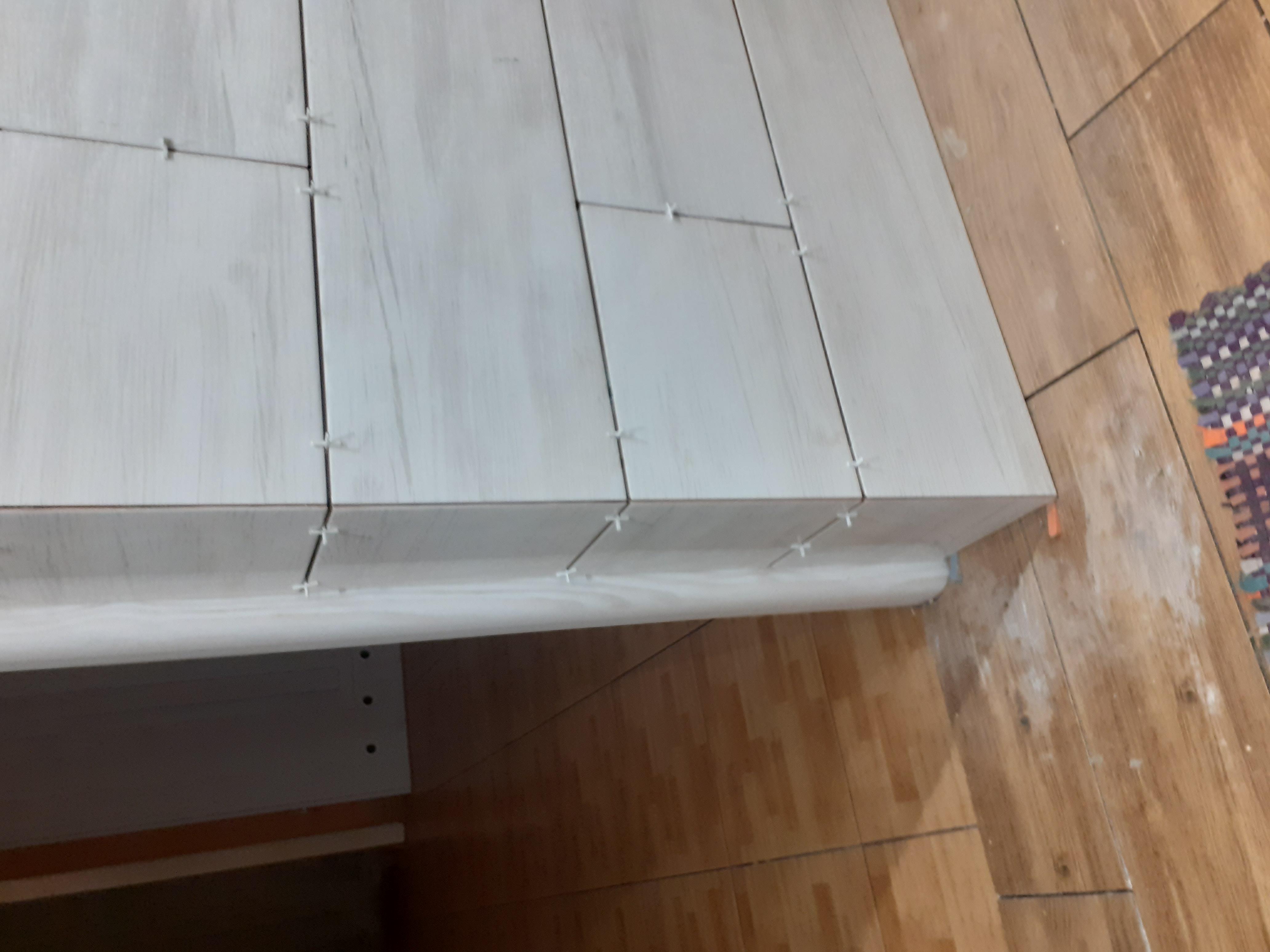 Świeżo ułożone, białe płytki podłogowe z drewnopodobnym wzorem, z plastikowymi krzyżykami dystansowymi w fugach, częściowo przykryte folią ochronną, widoczne na tle istniejącej podłogi z płytek...