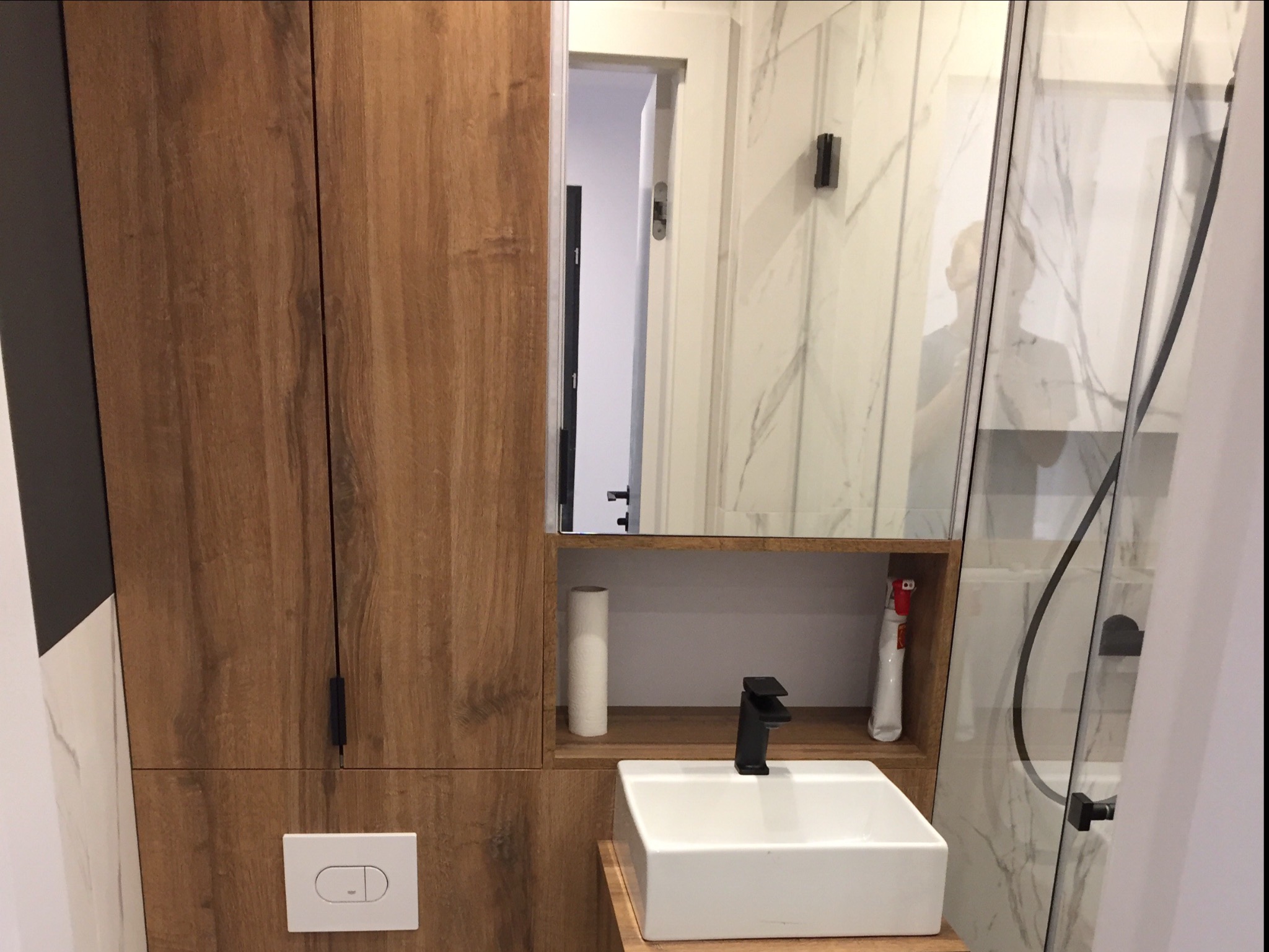 Nowoczesna łazienka z drewnianą zabudową, białą umywalką nablatową i kabiną prysznicową ze szklanymi drzwiami, widoczny fragment lustra z odbiciem osoby.