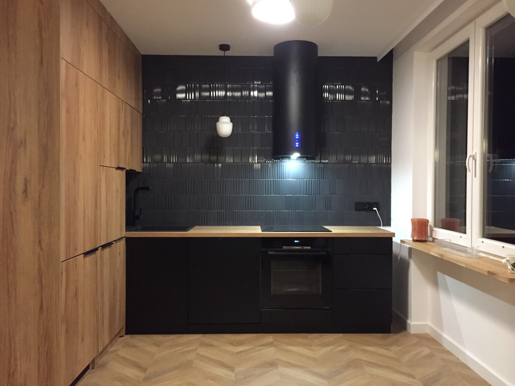 Nowoczesna kuchnia z czarnymi szafkami i drewnianymi frontami, czarny okap cylindryczny na tle ściany z ciemnych płytek, parkiet w jodełkę.