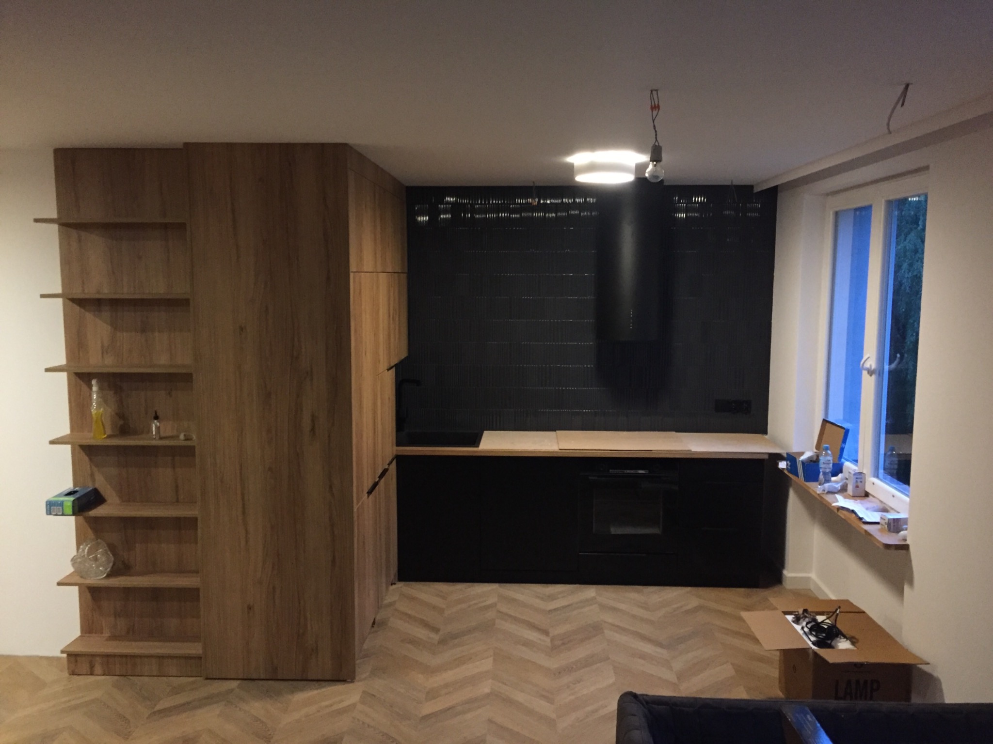 Nowoczesna kuchnia z drewnianymi szafkami, czarnymi frontami, czarną ścianą z płytek i parkietem w jodełkę, z kartonem po lampie na parapecie