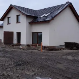 Dwukondygnacyjny dom jednorodzinny w trakcie budowy, z widoczną elewacją w kolorze złamanej bieli, ciemną dachówką i brązowymi ramami okiennymi, otoczony błotnistym terenem budowy z paletami...