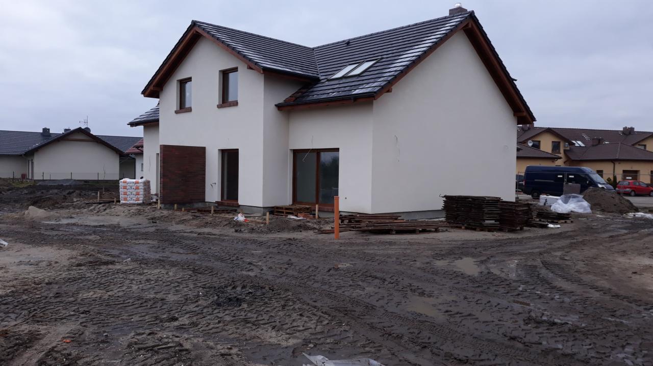 Dwukondygnacyjny dom jednorodzinny w trakcie budowy, z widoczną elewacją w kolorze złamanej bieli, ciemną dachówką i brązowymi ramami okiennymi, otoczony błotnistym terenem budowy z paletami...