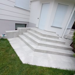 R&R Budownictwo - Schody wejściowe do domu w Wołominie wyłożone jasnymi płytkami ceramicznymi, z widocznymi fugami, prowadzące do białych drzwi z roletami.
