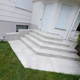 Schody wejściowe do domu w Wołominie wyłożone jasnymi płytkami ceramicznymi, z widocznymi fugami, prowadzące do białych drzwi z roletami.