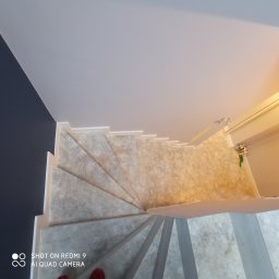 R&R Budownictwo - Wewnętrzne schody z szarymi stopniami i białymi podstopnicami, widok z góry, fragment buta w lewym dolnym rogu.