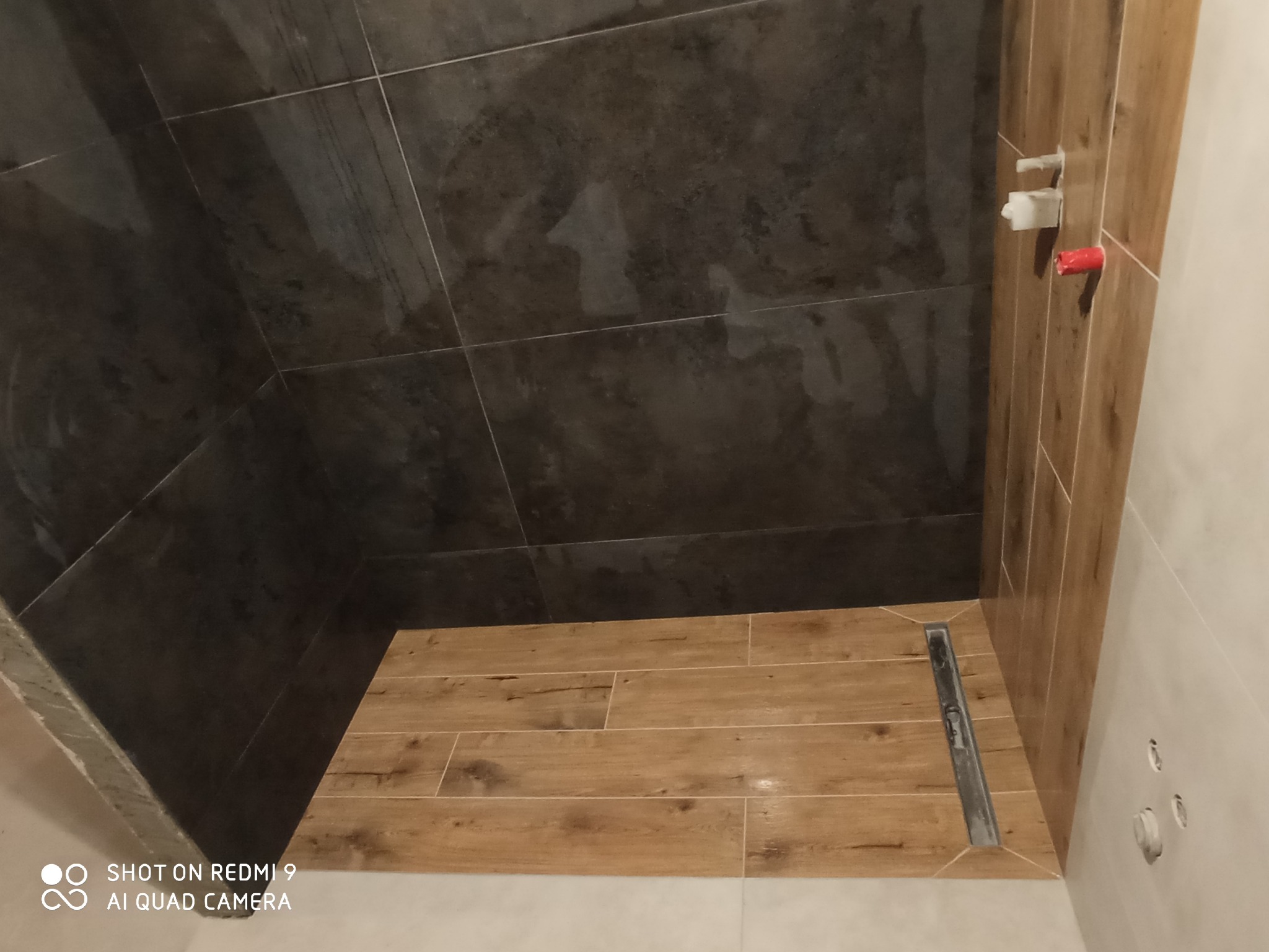 Ukończony fragment łazienki z ciemnymi płytkami na ścianach i drewnopodobnymi płytkami na podłodze, widoczny odpływ liniowy i fragment instalacji wodnej.