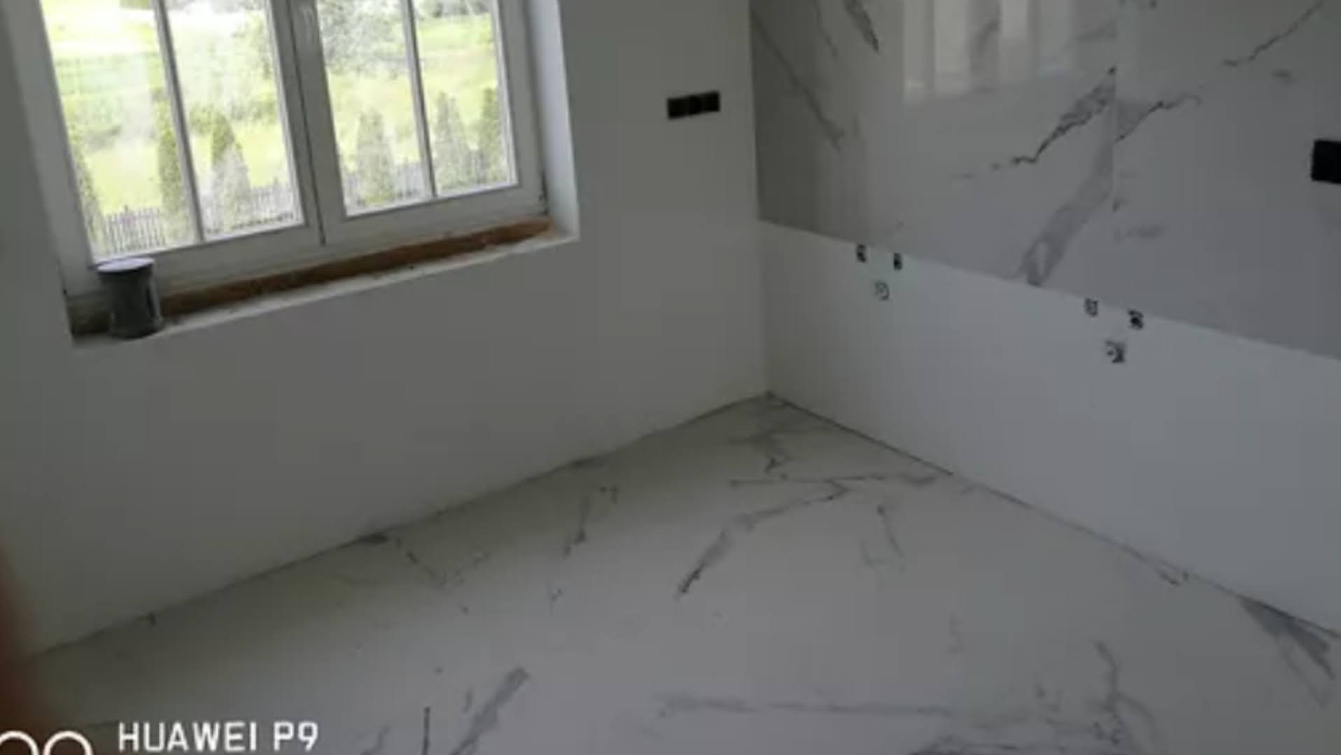 Wnętrze łazienki w trakcie prac wykończeniowych, widoczne białe płytki imitujące marmur na podłodze i częściowo na ścianach, przygotowane podłączenia elektryczne, puszka z farbą na parapecie okna.