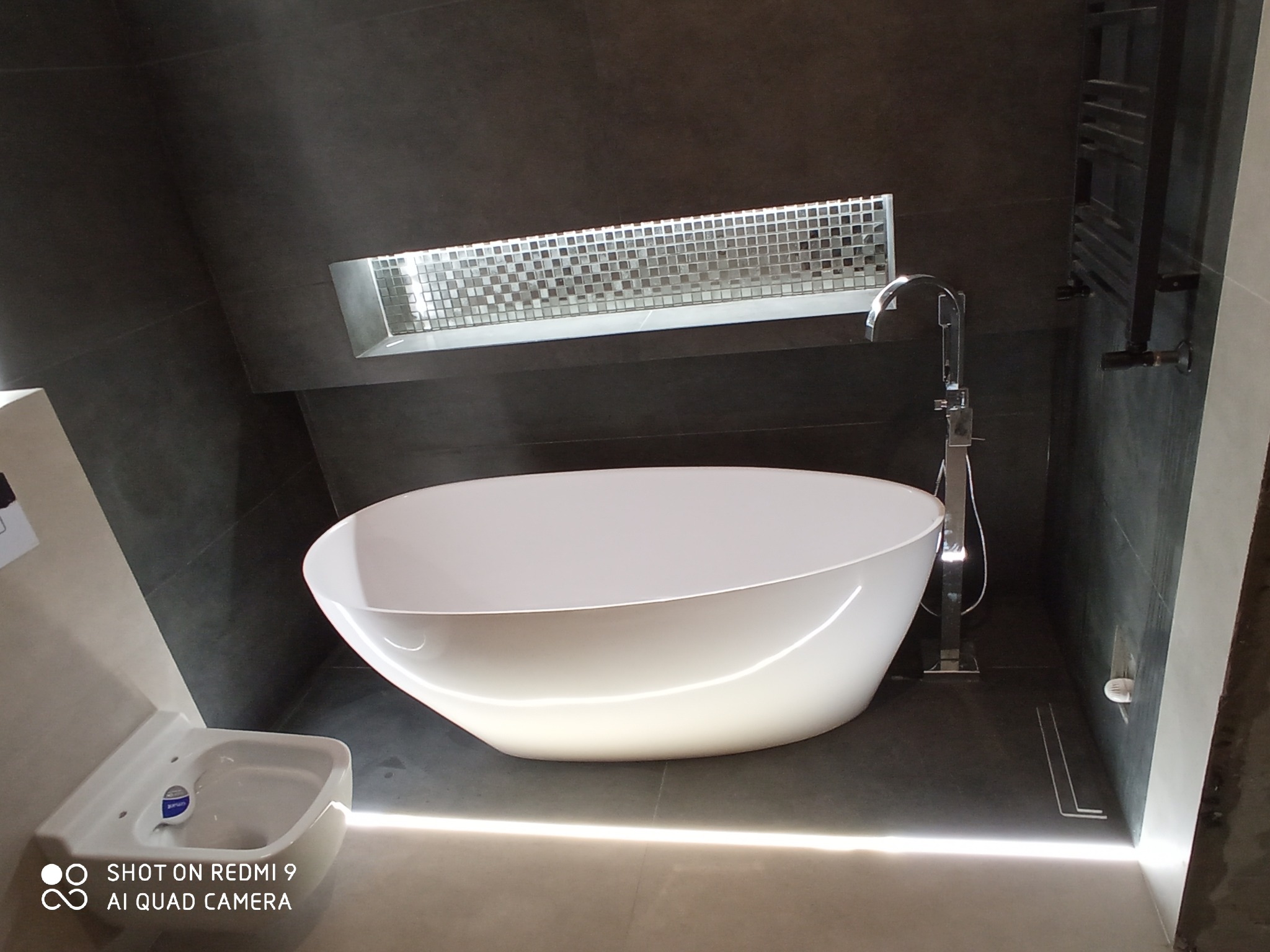 Nowoczesna łazienka z wolnostojącą białą wanną, armaturą w kolorze chromu i ciemnoszarą glazurą, oświetlona listwą LED pod wanną i mozaiką w niszy ściennej.