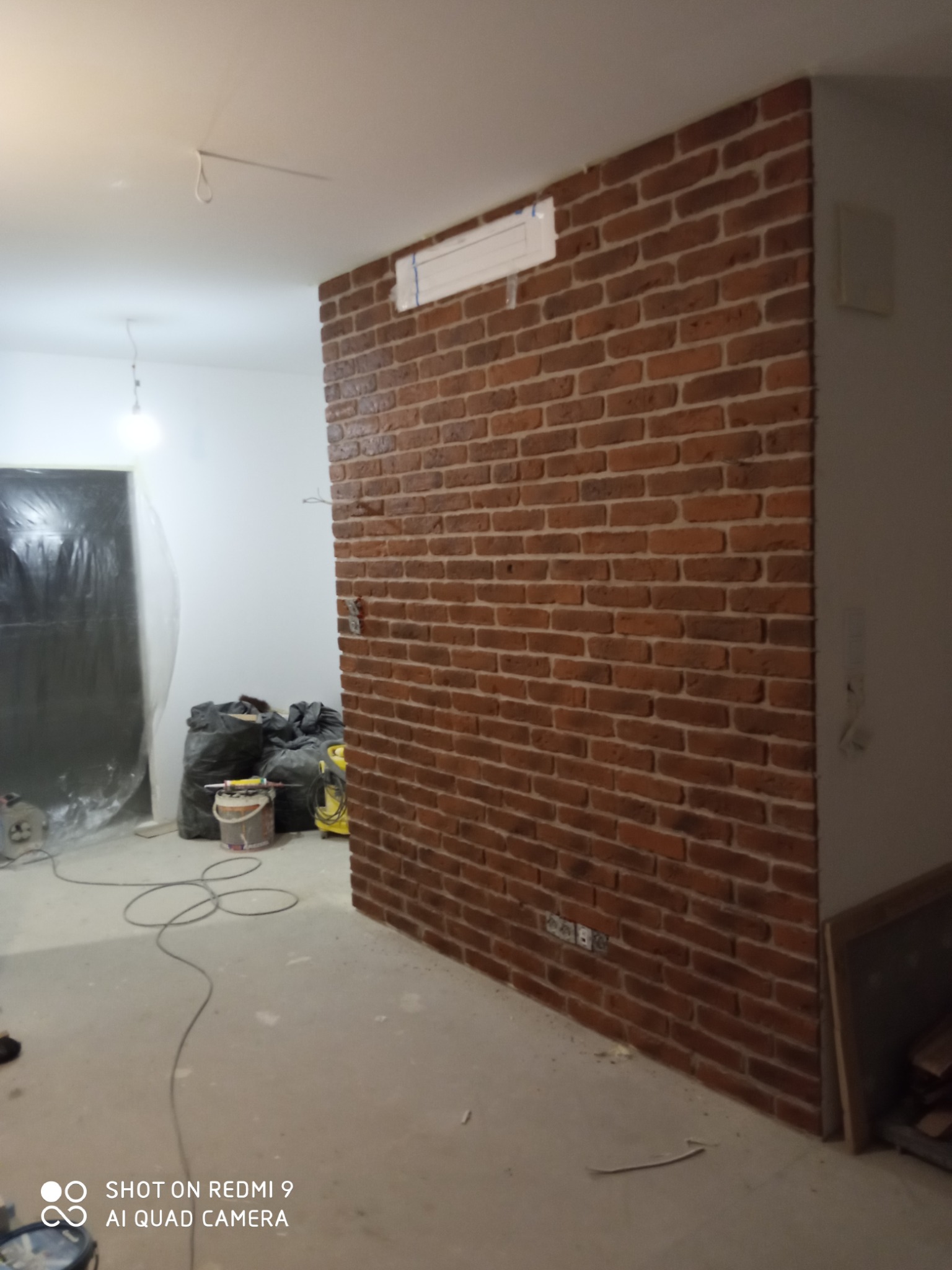Ściana wykończona czerwoną cegłą klinkierową, widoczne gniazdka elektryczne, worki z resztkami budowlanymi i kable na podłodze, surowy stan wykończenia.