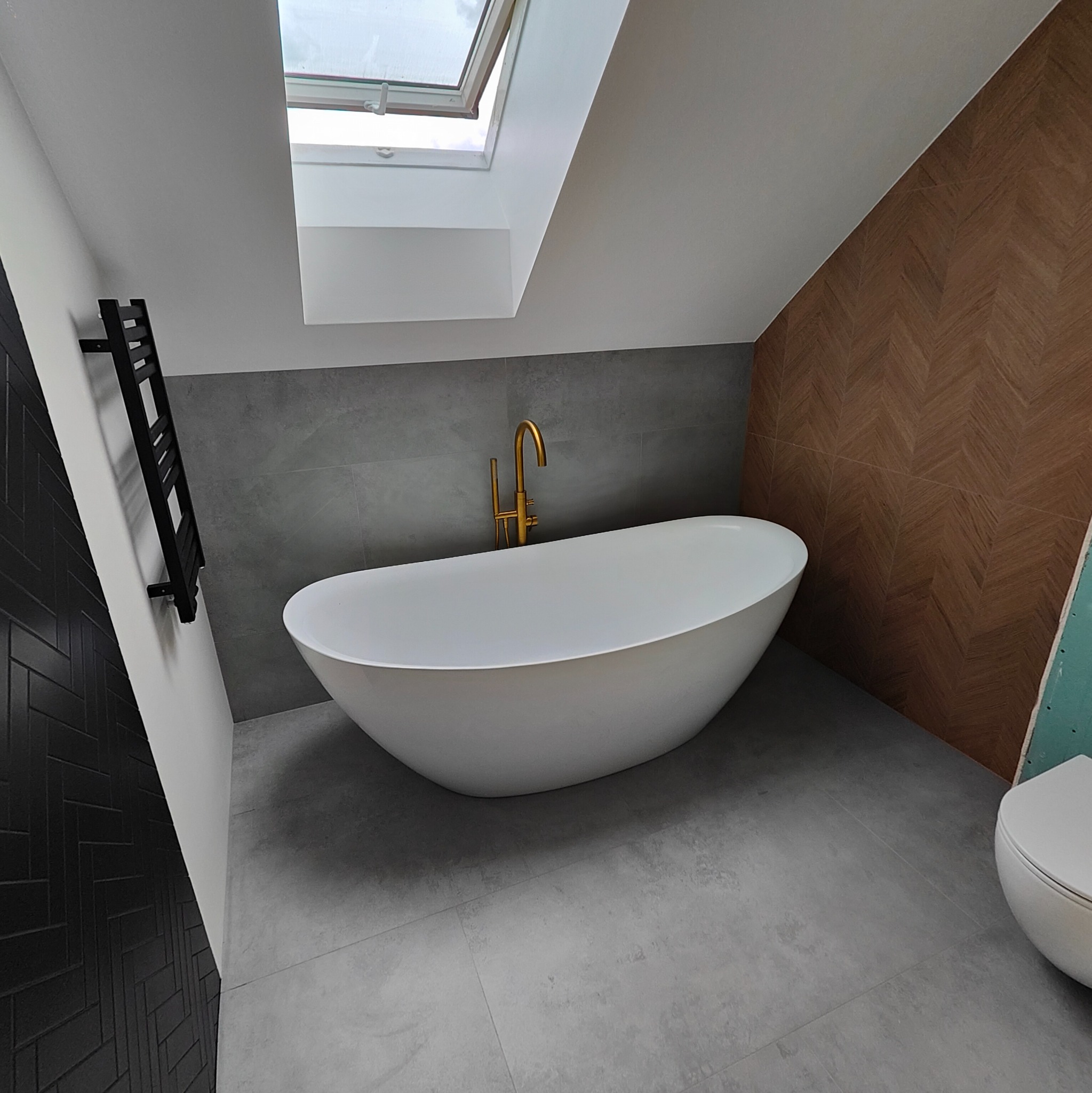 Nowoczesna łazienka na poddaszu z wolnostojącą białą wanną, złotą armaturą, szarą podłogą i ścianami z płytek oraz drewnianą ścianą w jodełkę, doświetlona oknem dachowym.