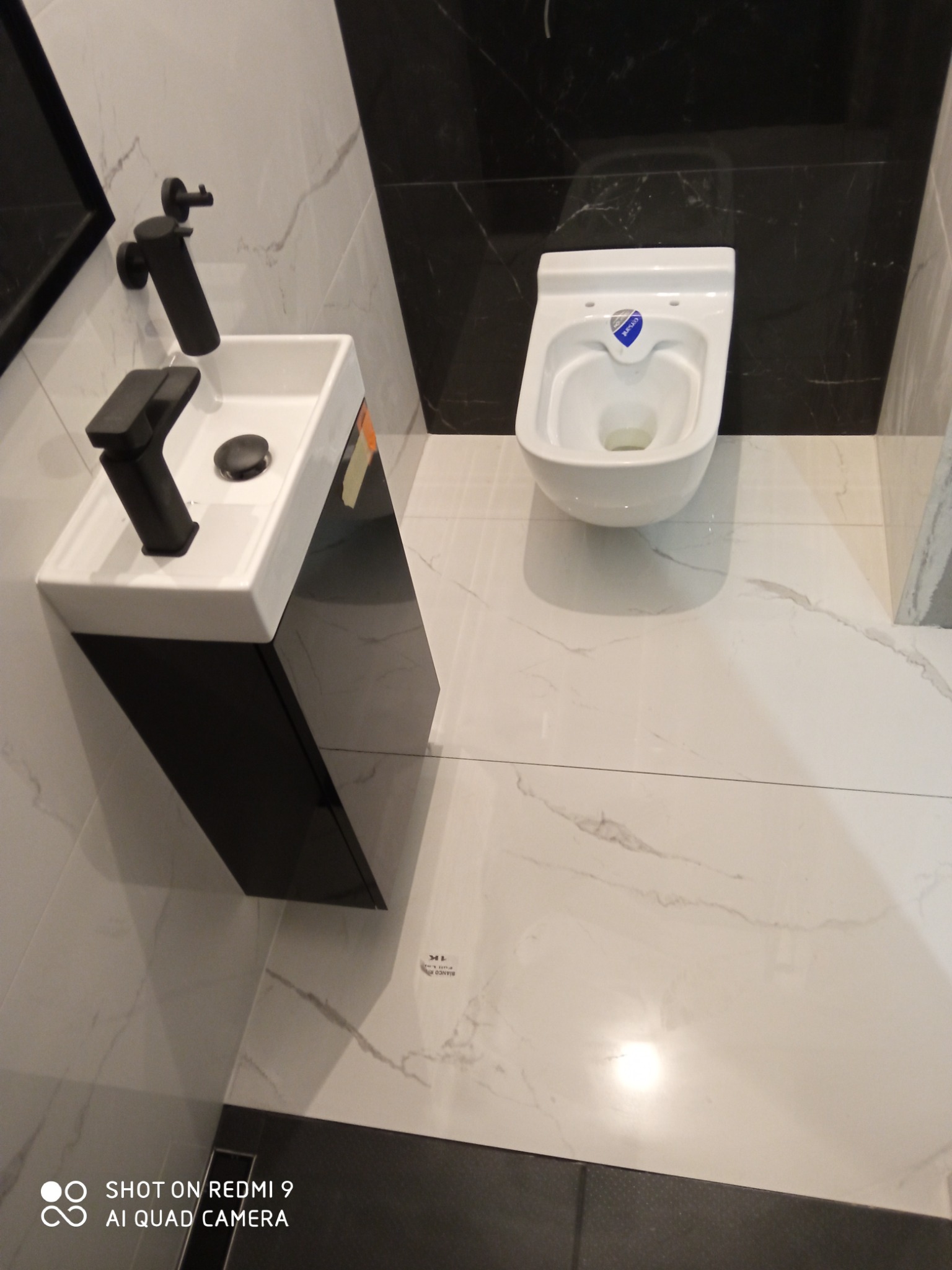 Nowoczesna łazienka z biało-czarnymi płytkami imitującymi marmur, wisząca toaleta z niebieskim logo, mała umywalka z czarną szafką i baterią.