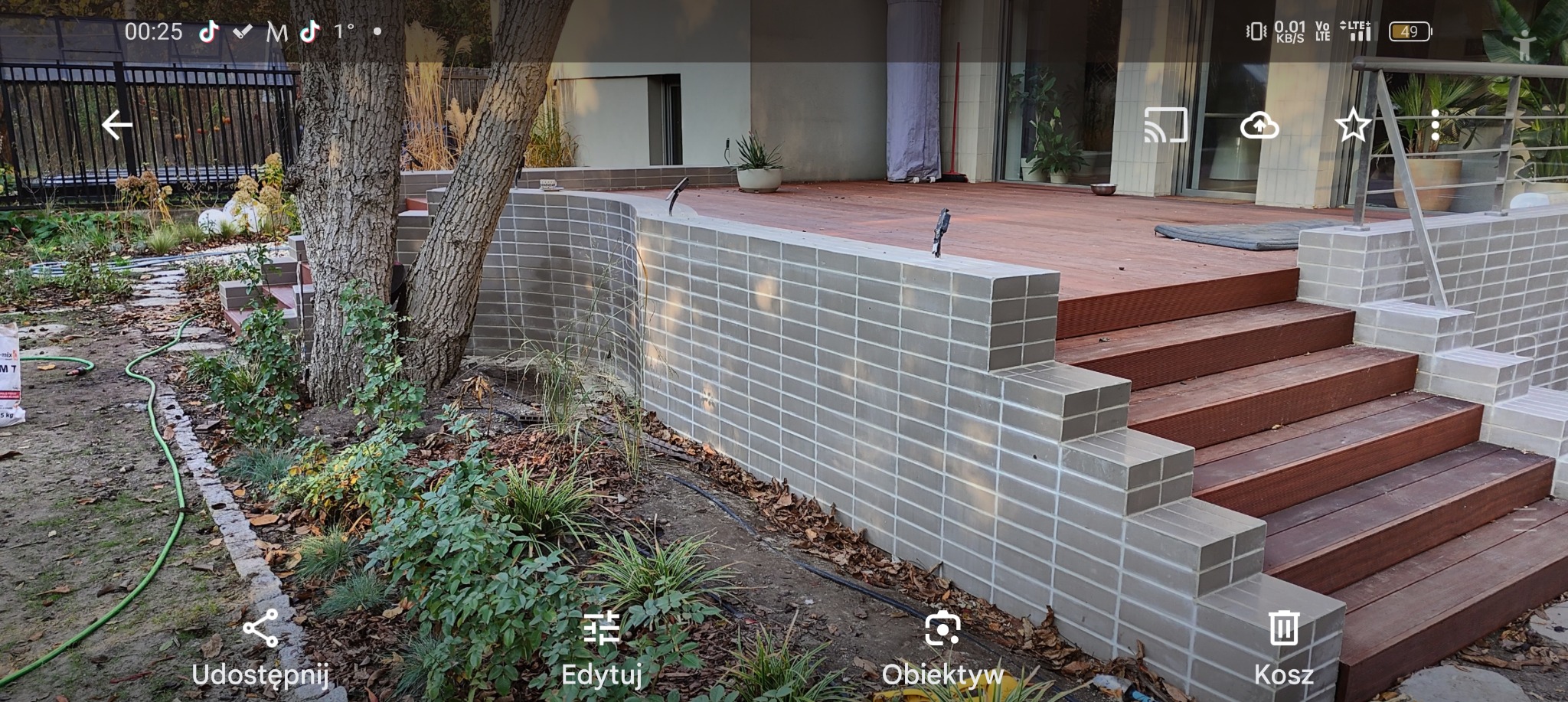 Nowoczesny taras z drewnianymi schodami i murem z szarych płytek, otoczony zielenią i drzewami.