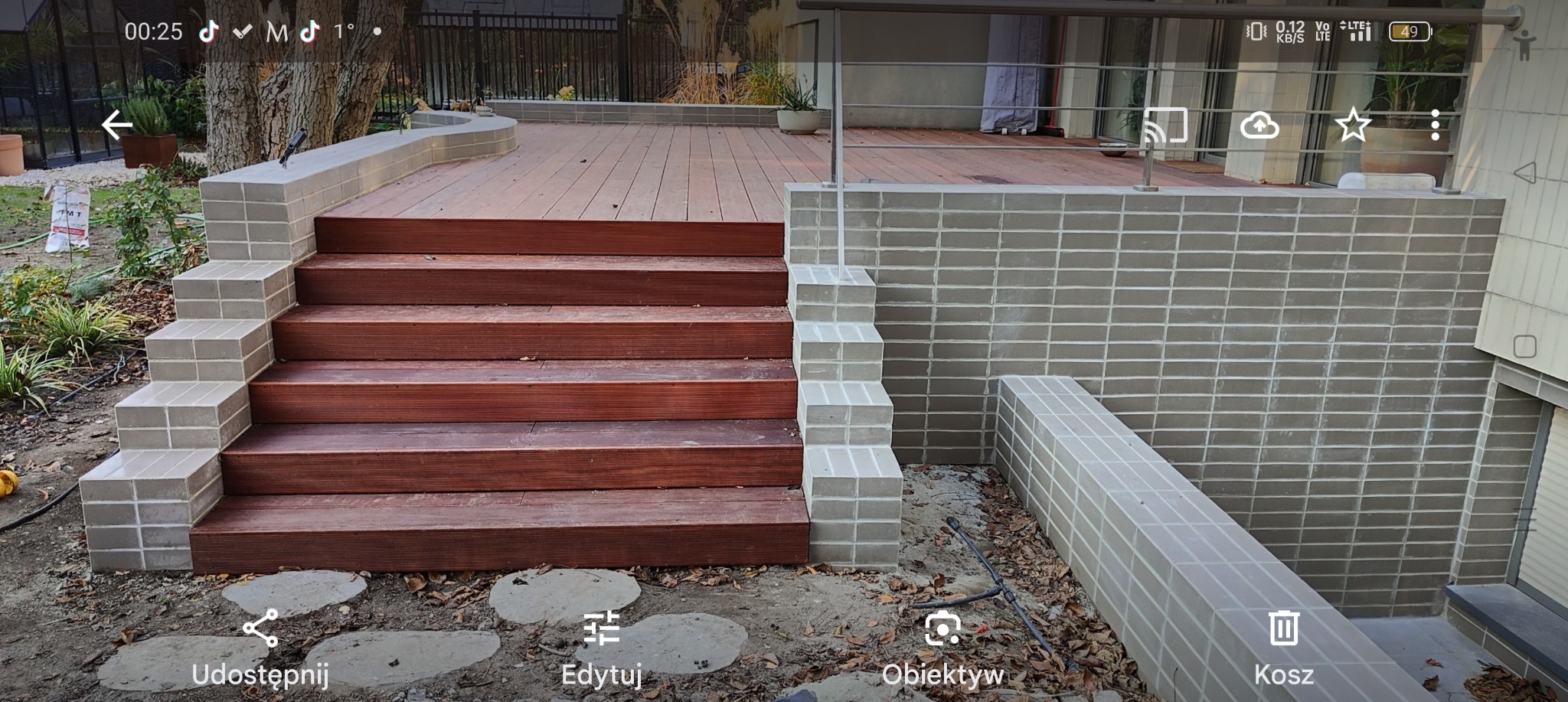 Schody z drewna kompozytowego prowadzące na taras, obłożone cegłą klinkierową w odcieniach szarości, z widocznymi śladami montażu w postaci ziemi i liści.