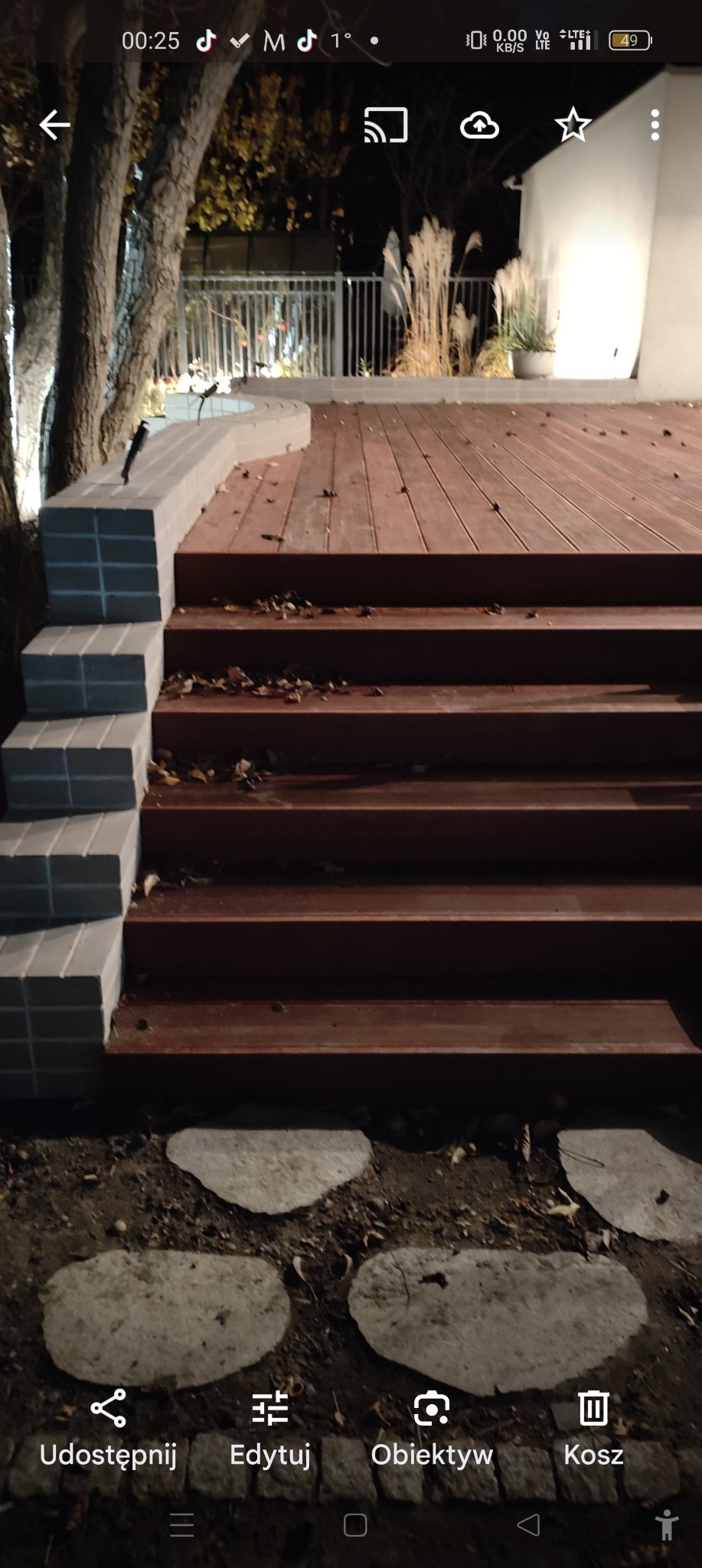 Schody z drewnianymi stopniami i obłożone szarą kostką, prowadzące na drewniany taras z metalową barierką, oświetlone nocą