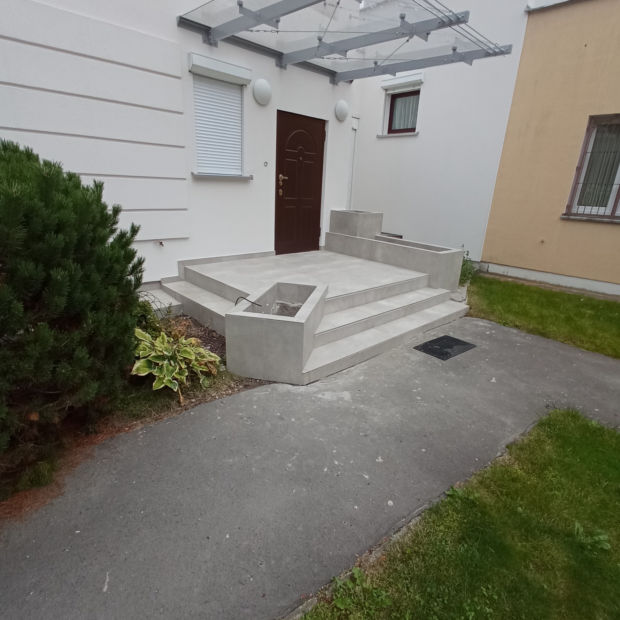 Wykończone schody wejściowe do budynku z szarych płytek, z donicami z tego samego materiału, widok z zewnątrz.