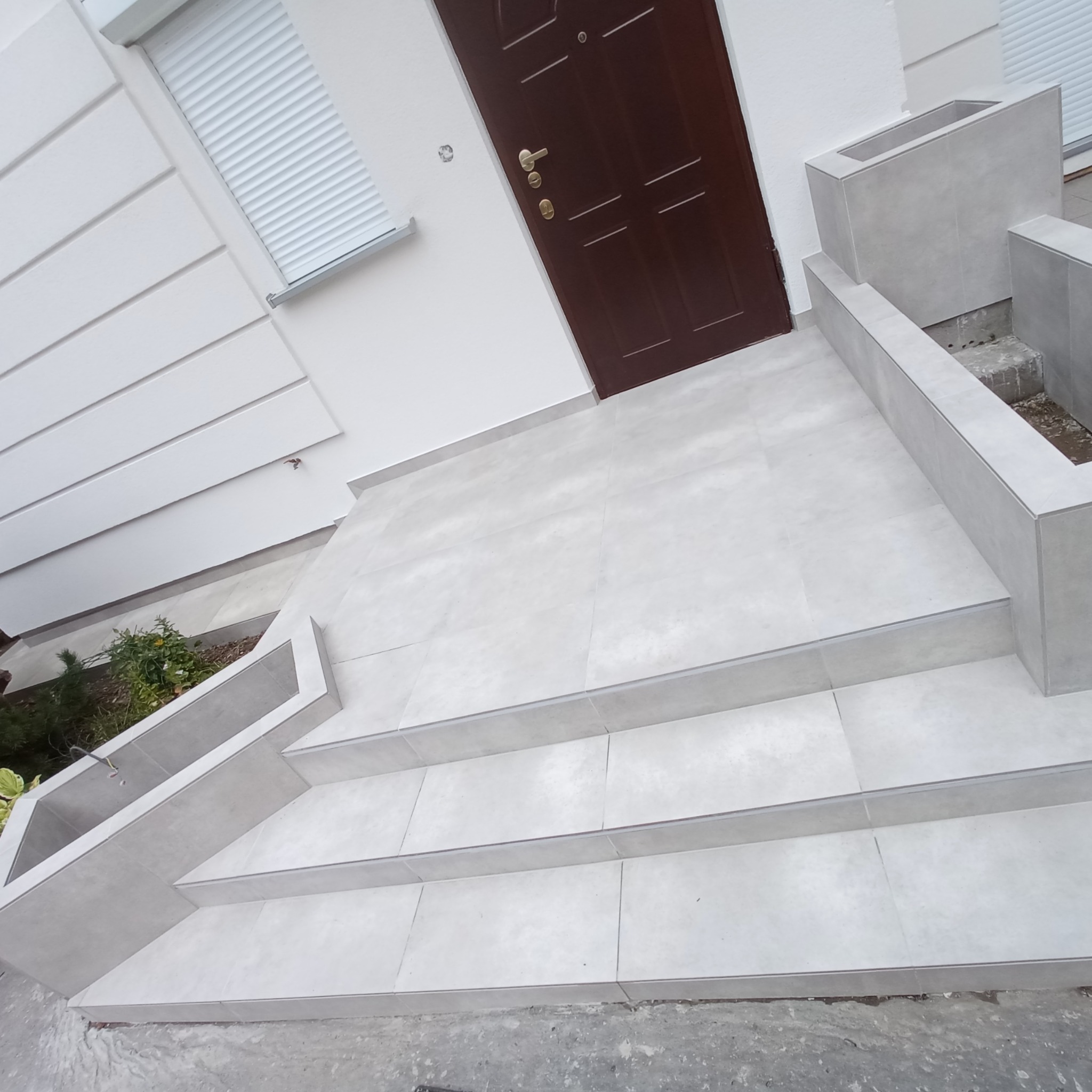 Wyłożone jasnoszarymi płytkami schody wejściowe z donicami, brązowe drzwi wejściowe, biała elewacja z żaluzjami.