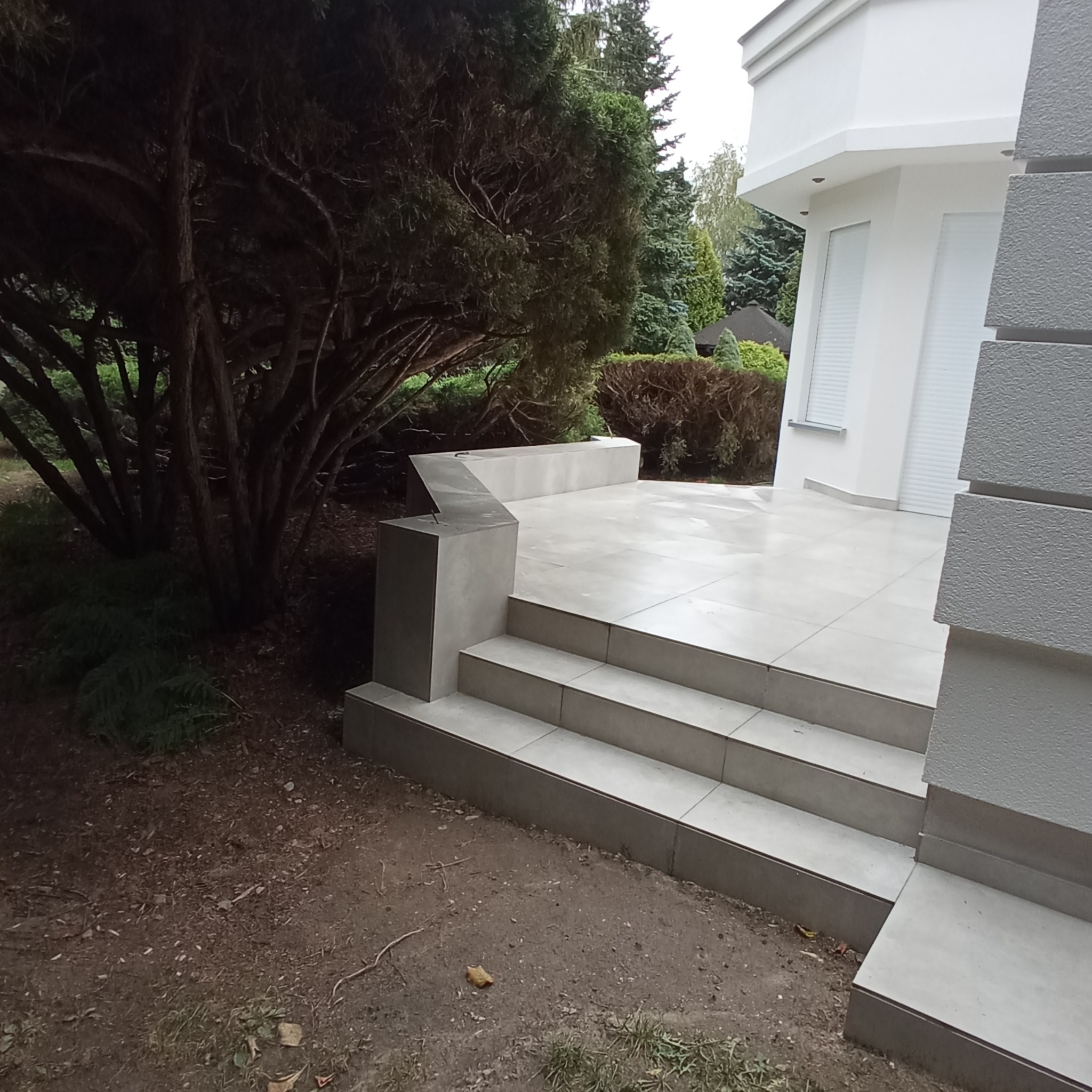 Wyłożony jasnymi płytkami taras z trzema schodami, otoczony zielenią i elementami nowoczesnej architektury.