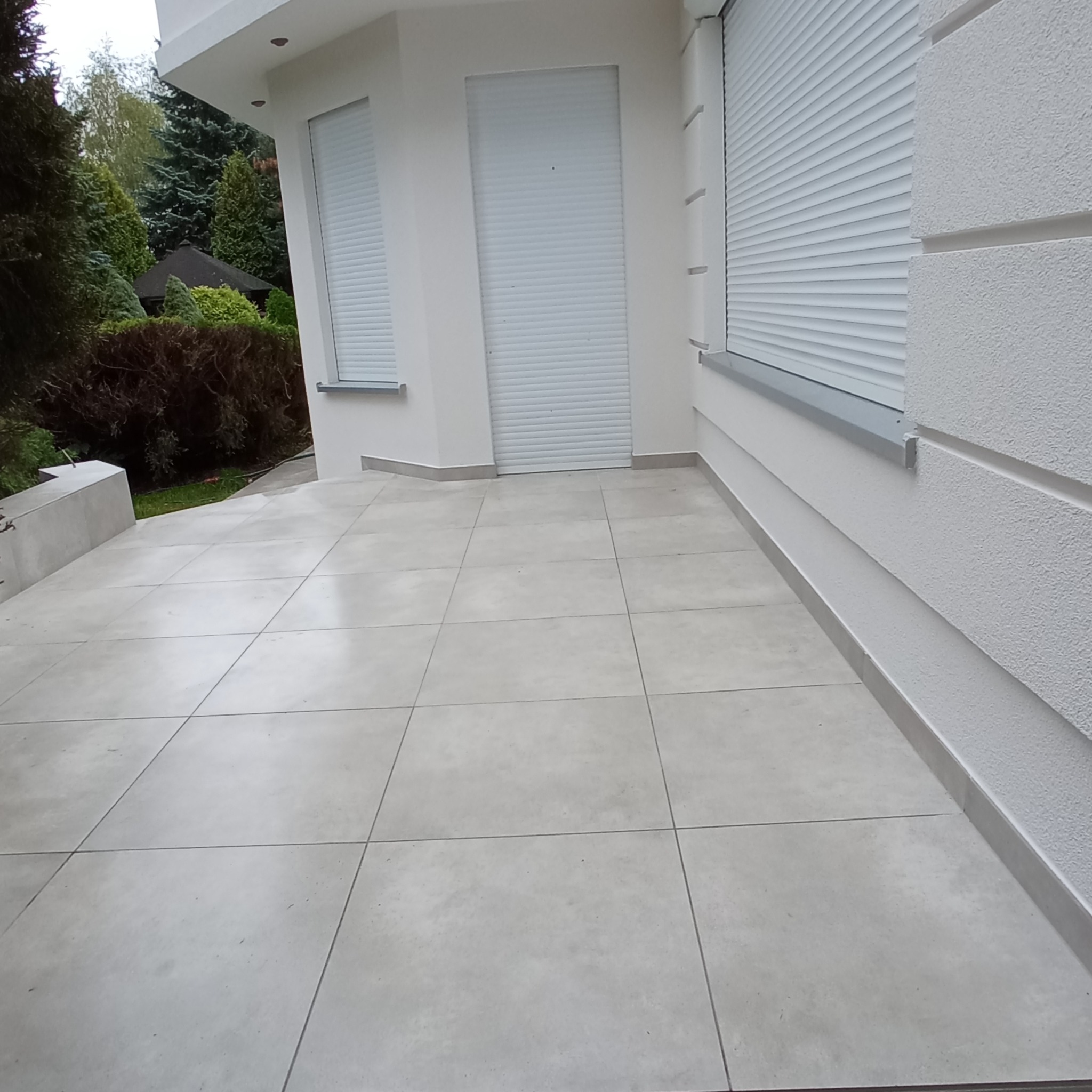 Wyłożony jasnoszarymi płytkami taras przylegający do białego budynku z zamkniętymi roletami okiennymi.