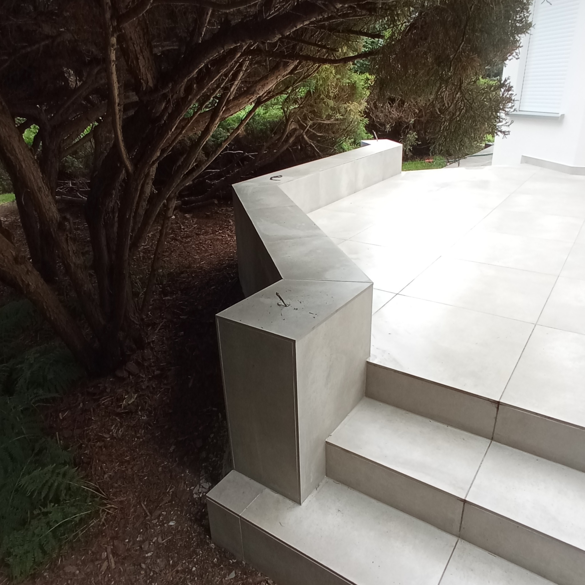 Wyłożone jasnymi płytkami schody zewnętrzne z betonową balustradą, widoczne drzewo iglaste w tle.