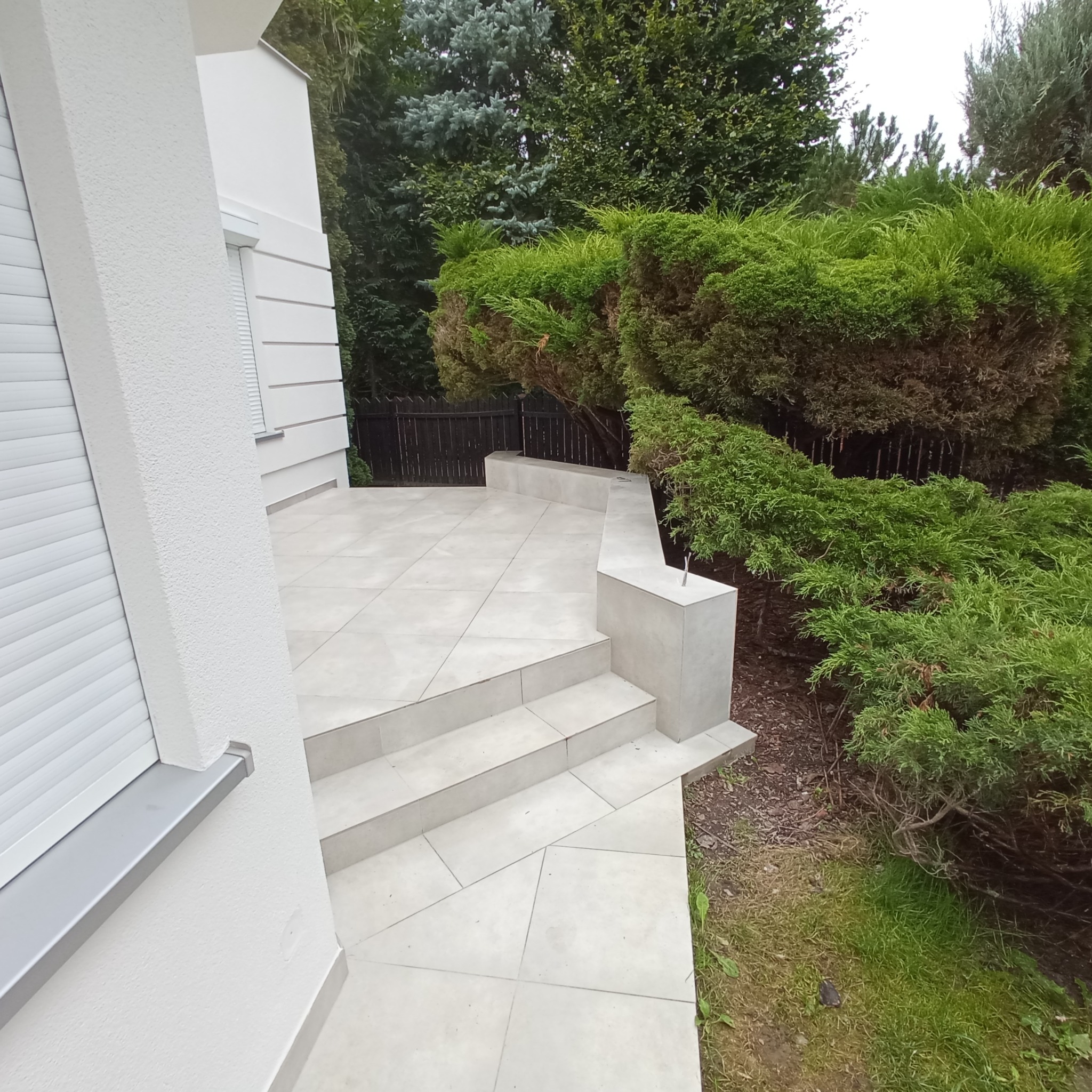 Jasnoszara, gładka, ceramiczna podłoga tarasu z trzema schodkami, otoczona zielenią, przylegająca do białego budynku z roletami zewnętrznymi.