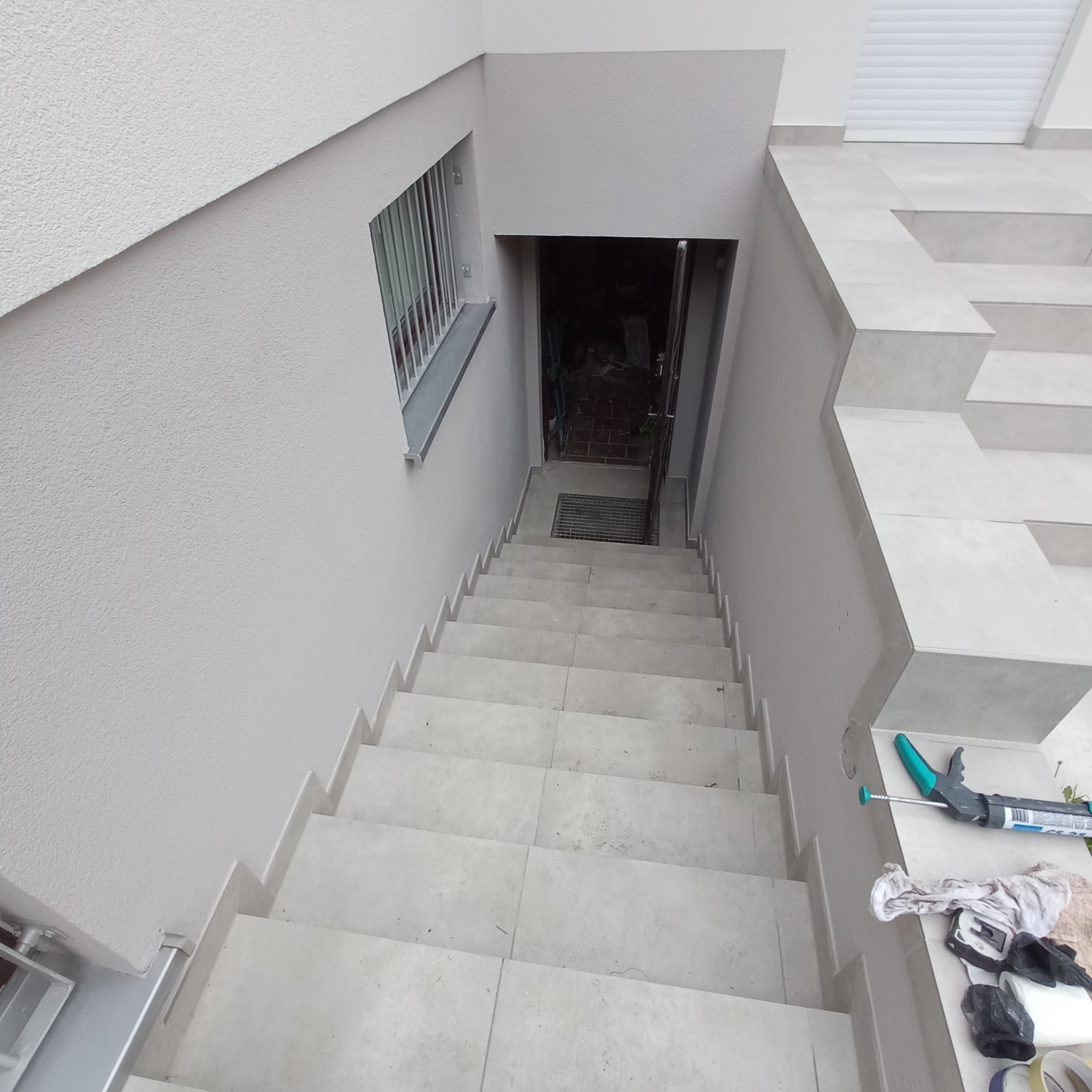 Wykończone płytkami schody zewnętrzne prowadzące do piwnicy, widok z góry, na dole widoczne otwarte drzwi, na górze okno z kratą, na jednym ze stopni leżą narzędzia glazurnicze.