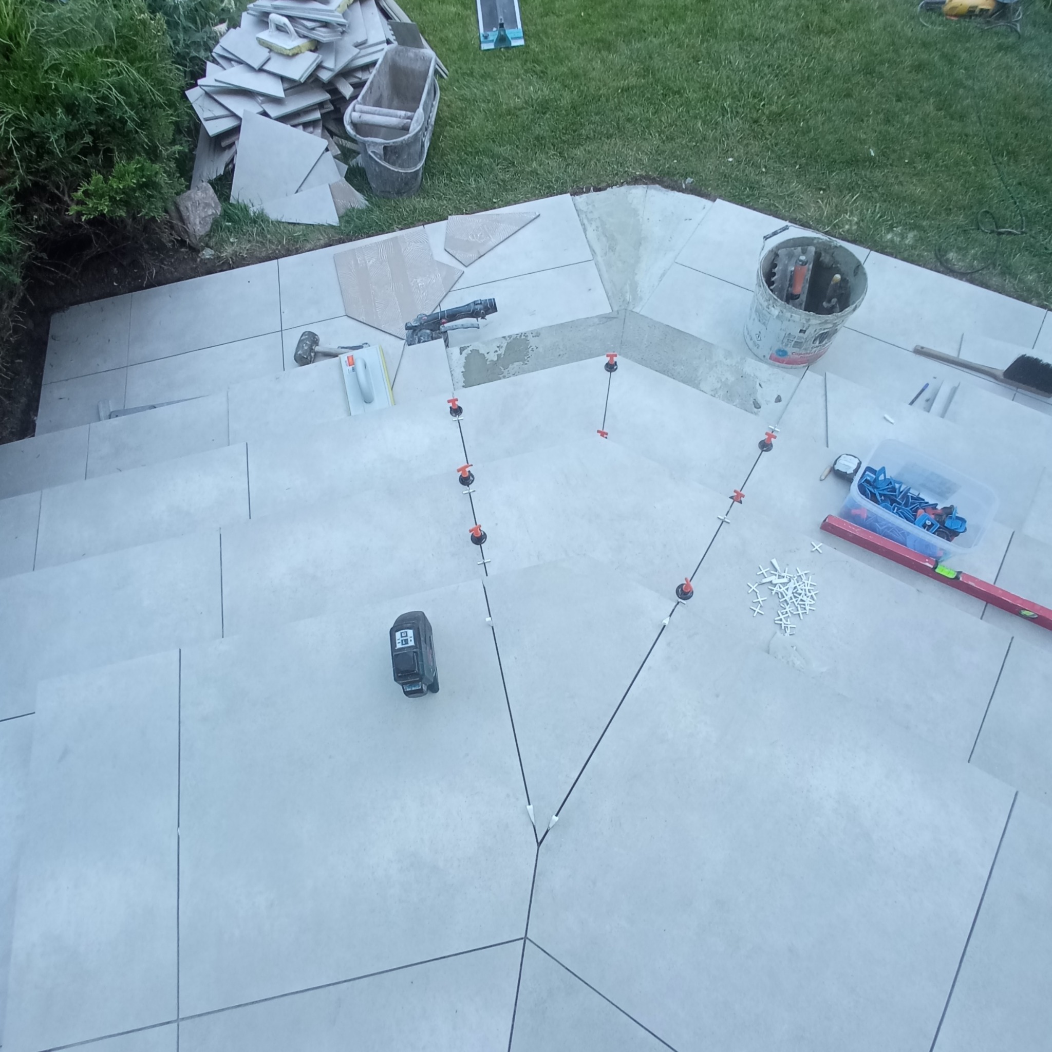 Układanie jasnoszarych płytek na schodach zewnętrznych, widoczne narzędzia glazurnicze: poziomica laserowa, system poziomowania płytek, zapas płytek, wiadro z zaprawą.