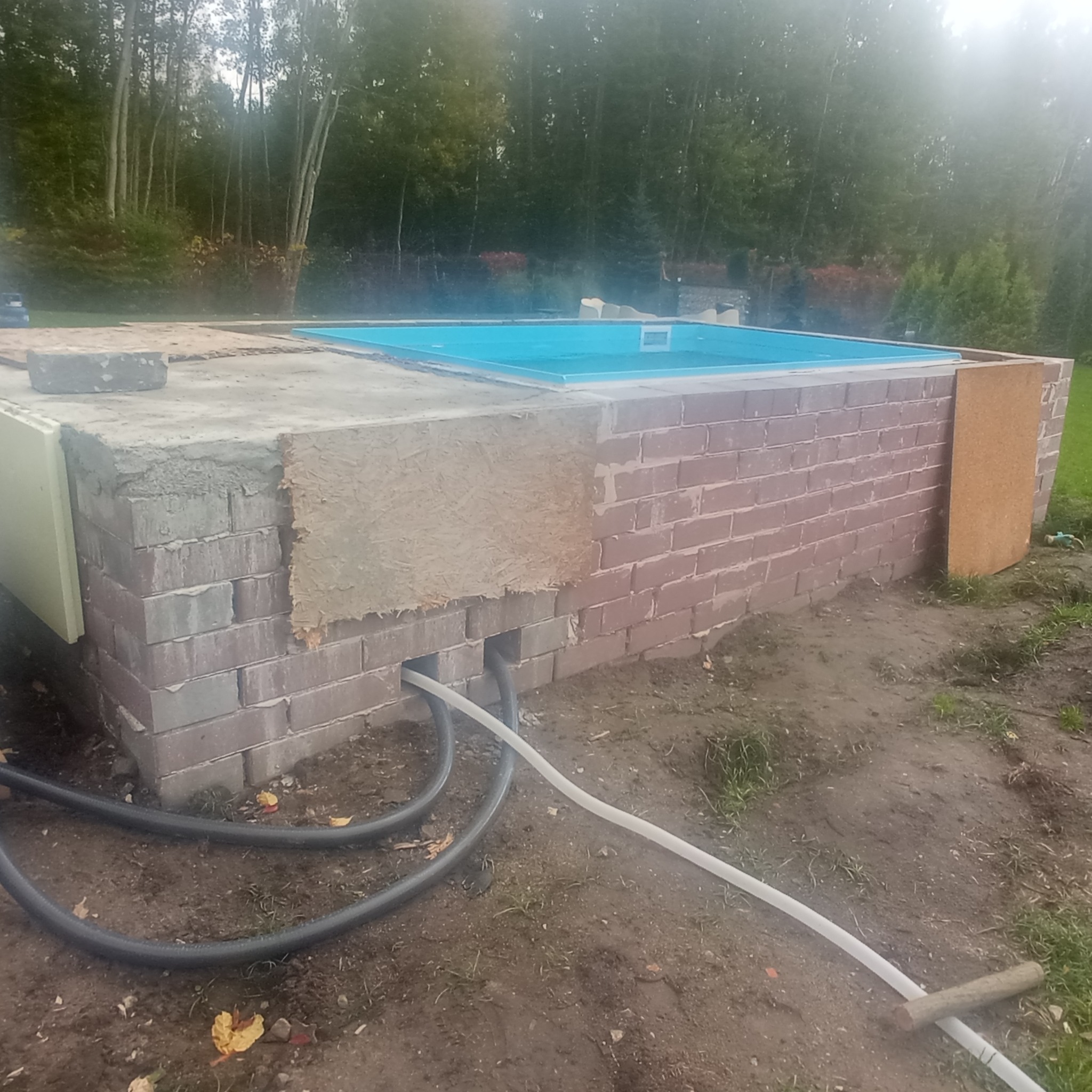 Budowa zewnętrznego basenu: murowane ściany z cegły, częściowo otynkowane, z widocznymi przewodami i niebieskim wnętrzem basenu w surowym otoczeniu.