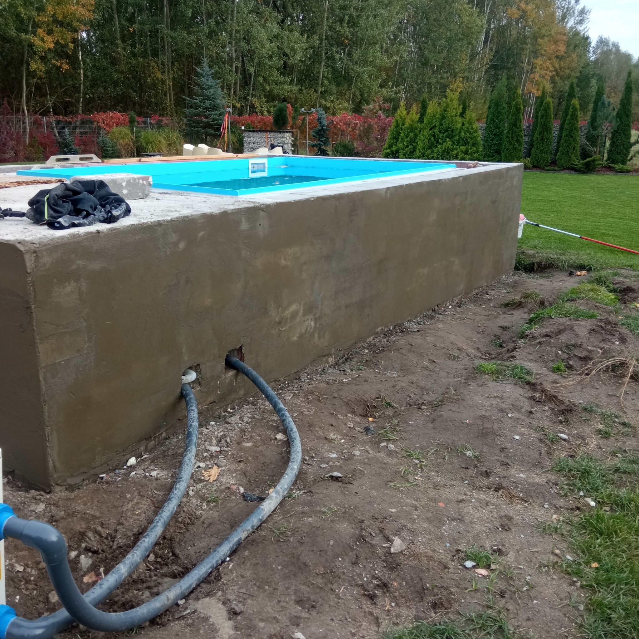 Betonowa konstrukcja basenu w trakcie budowy, z widocznymi rurami instalacji, na tle zieleni ogrodowej.