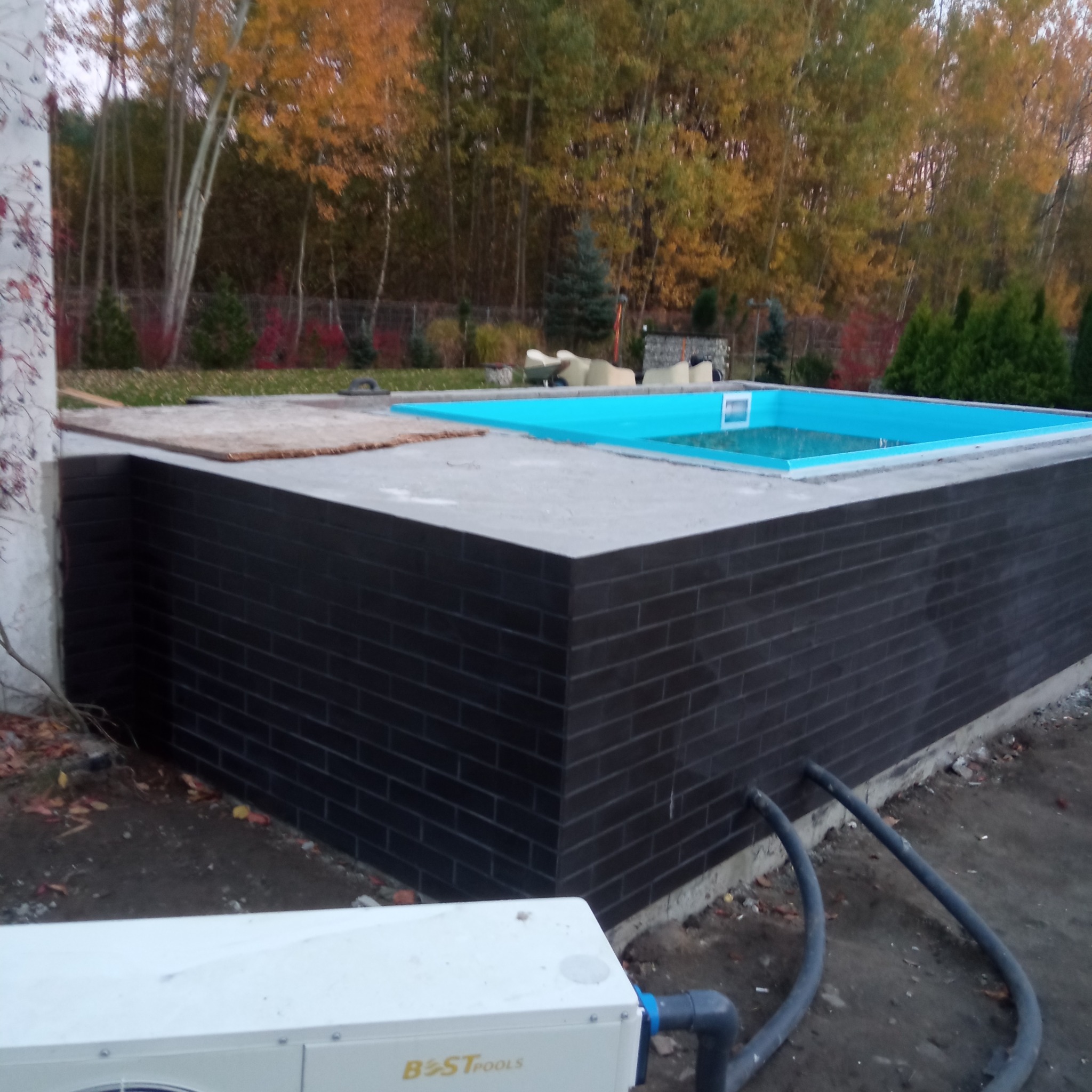 Budowa basenu zewnętrznego z ciemną, ceglaną elewacją i niebieską folią w tle jesiennego ogrodu.